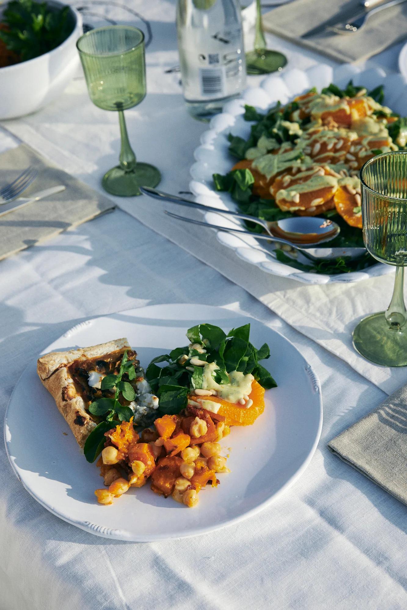 Outdoor table with vegetarian pizza, salad, and chickpea dish. Green wine glasses and utensils visible.