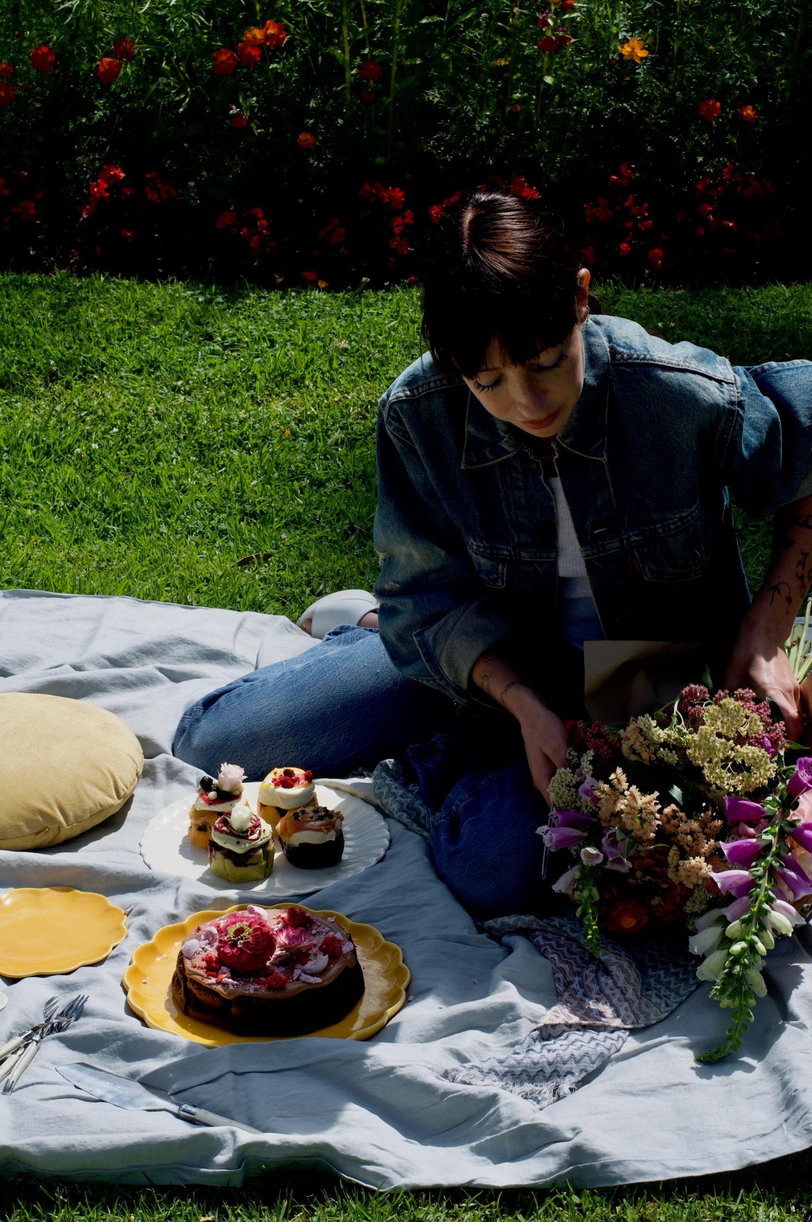 A person in a denim jacket arranges flowers on a picnic blanket with desserts on yellow plates. Bright flowers bloom nearby, evoking The Caker’s Auckland bakery vibe.