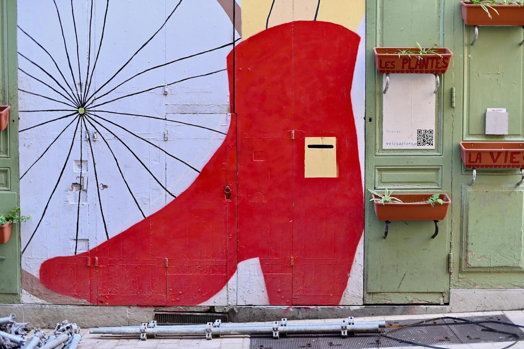 Mural of large red boot on street with green doors. Doors have small planters labeled "Les Plantes" and "La Vie".