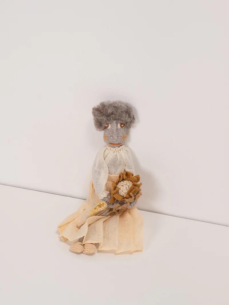 A hand-crafted doll with gray hair, a beige dress, and a small bouquet, seated against a white background. The intricate details reflect Sophie Digard’s craftsmanship.