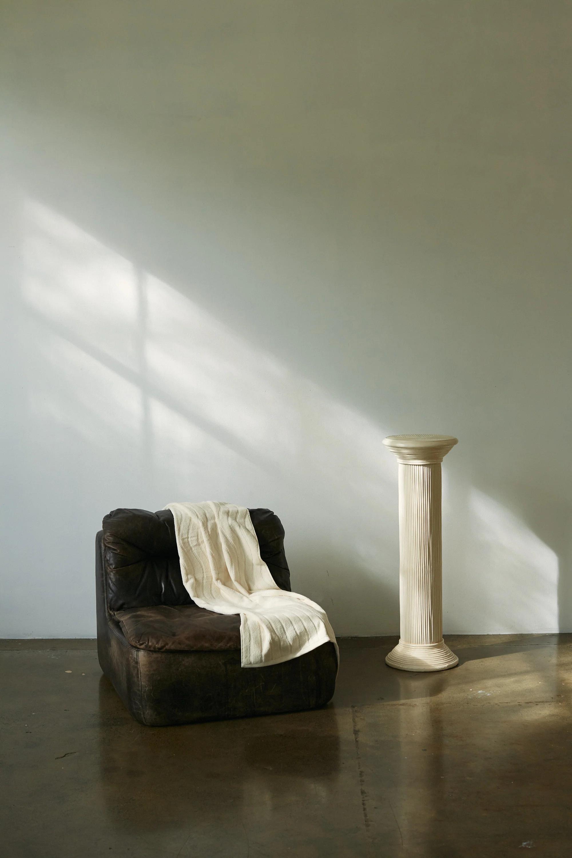 A minimalist interior with a dark leather armchair draped with a white textile, next to a white fluted pedestal. Soft natural light casts shadows, creating a serene, timeless space.