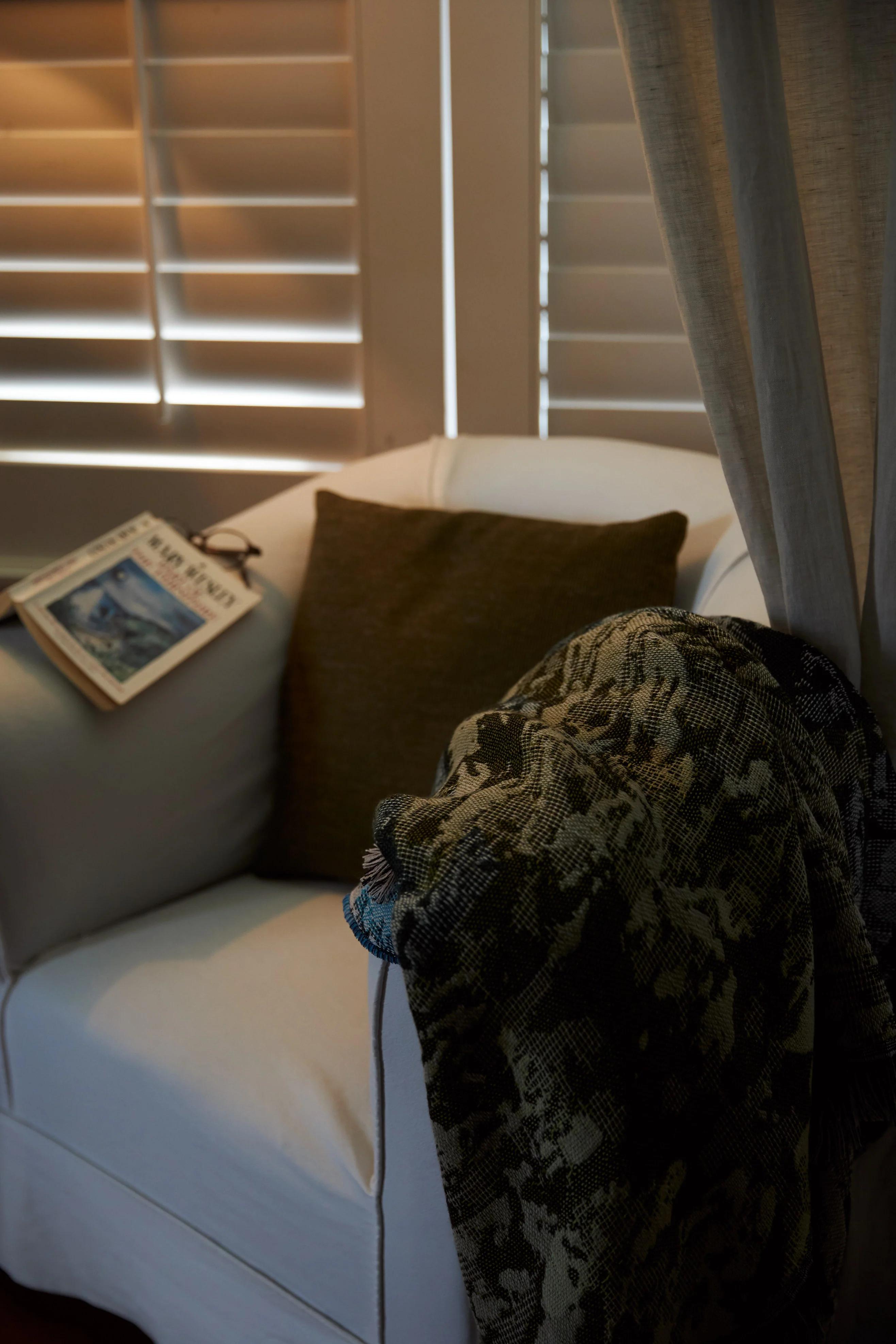 A white armchair with a brown cushion and draped blanket sits by closed shutters, with a book and glasses resting on the arm, creating a comfortable bedroom space.