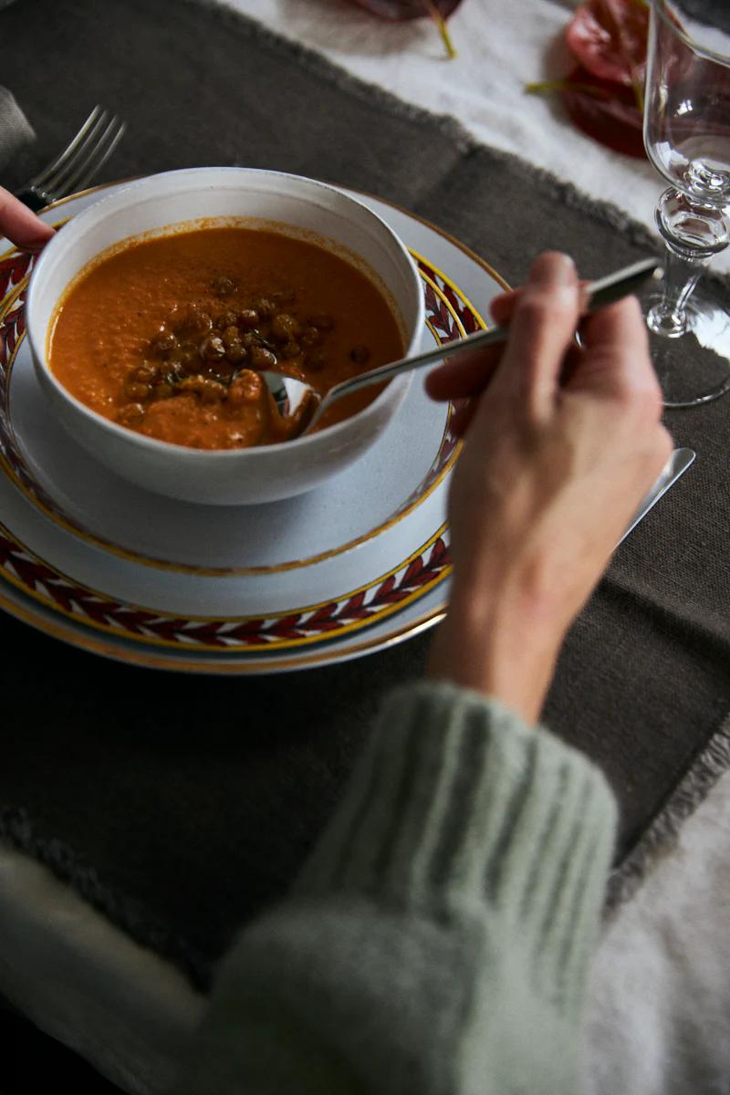 A person in green enjoys tomato and lentil soup from a white bowl. The table is set with a placemat, napkin, and wine glass.