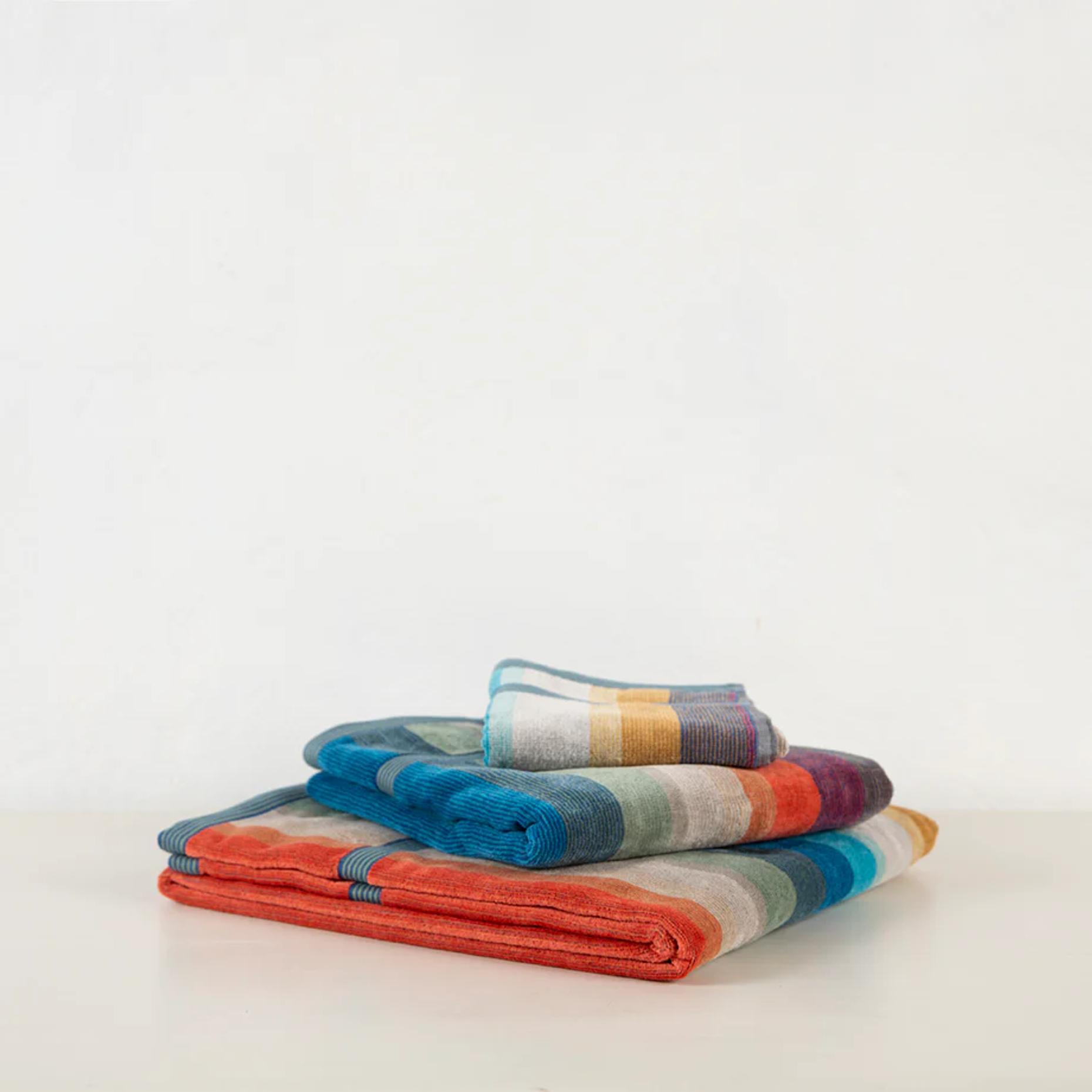 Four folded Missoni towels stacked on white surface. Vibrant stripes in orange, blue, green, yellow, and red showcase intricate textile design.