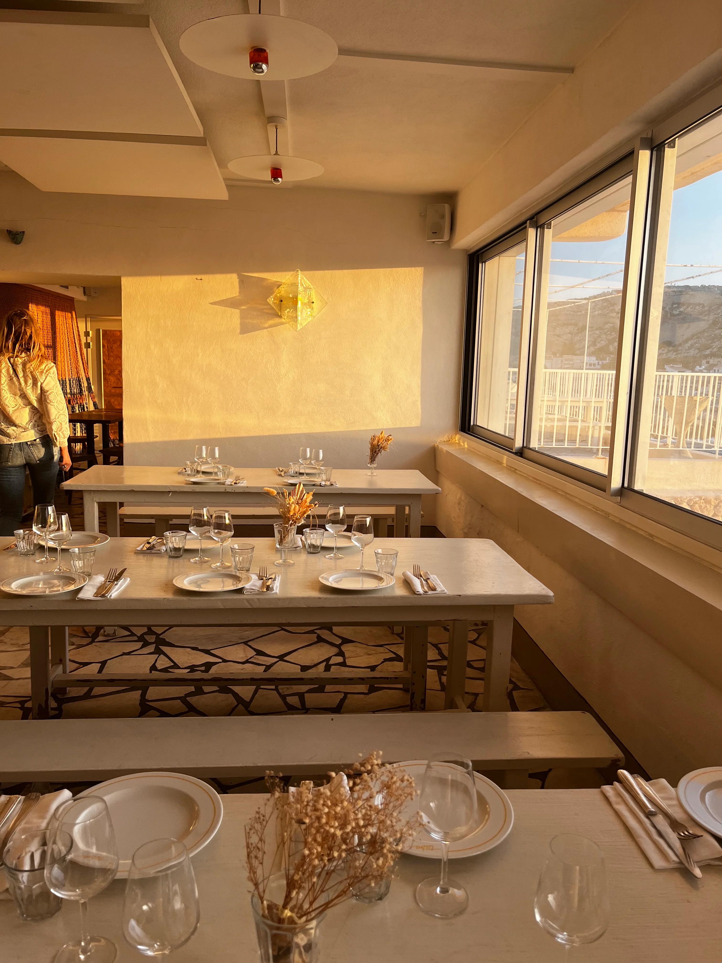 Cozy restaurant interior with neatly set tables and dried floral arrangements. Large windows offer scenic views. A person stands near the entrance.