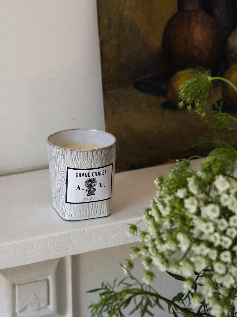 White "Grand Chalet - A.V. Paris" candle on ledge near green plant. Blurred dark painting in background.