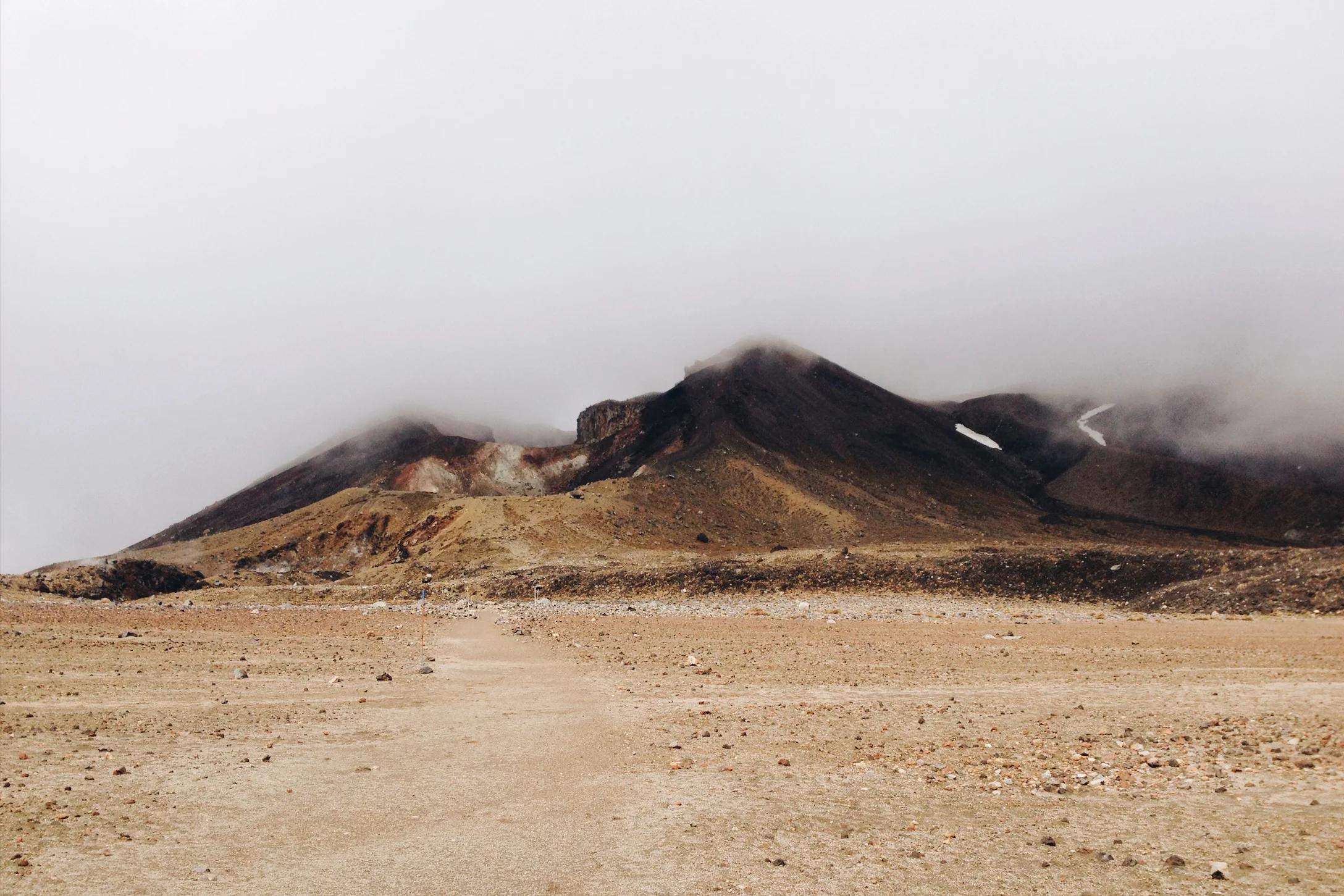 Barren, rocky landscape leading to misty mountains. Trail stretches to mountain base. Overcast sky and fog-obscured peaks create serene yet eerie atmosphere in untouched country.