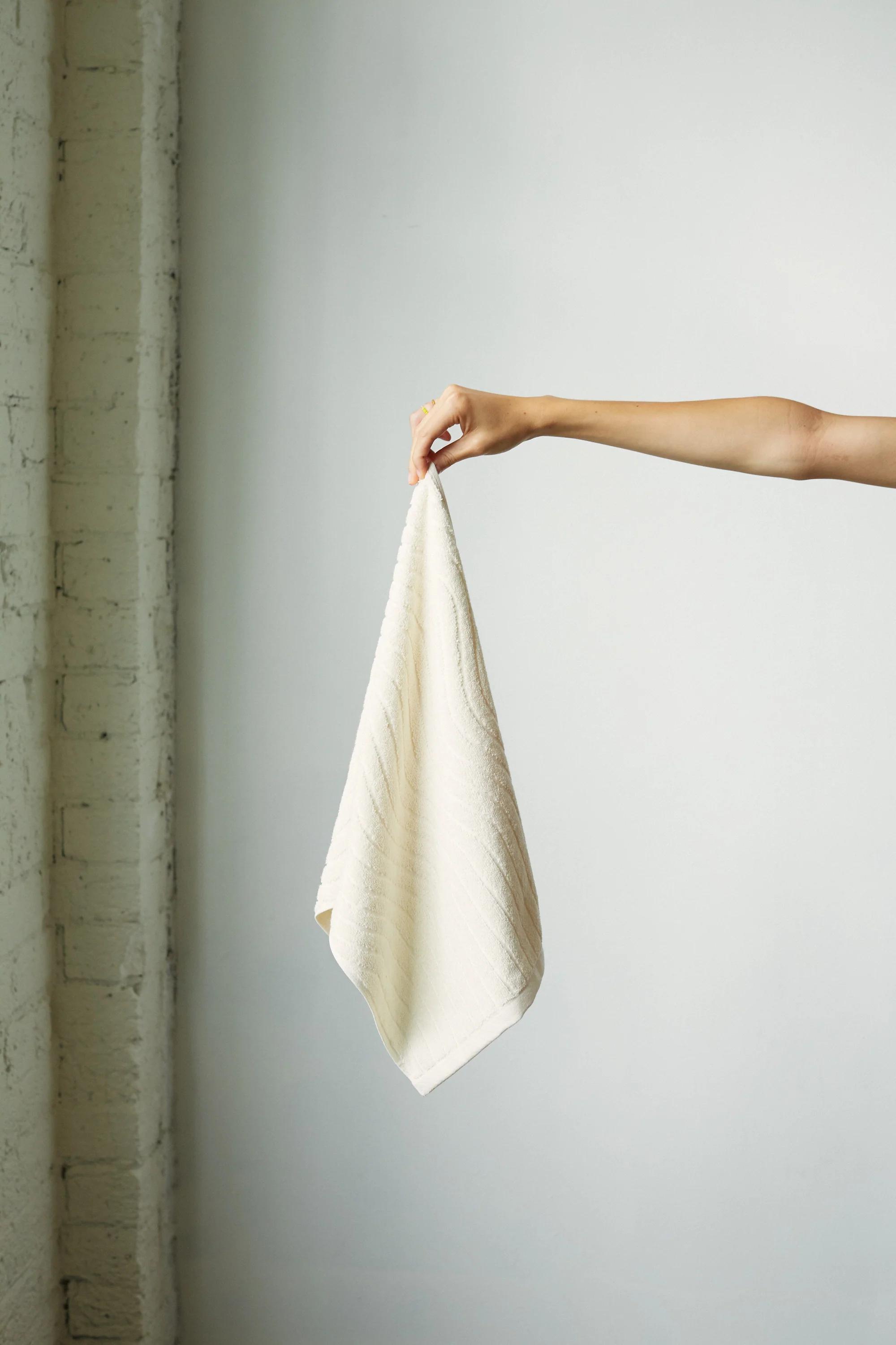 A person’s arm extends from the right, holding a light beige towel by one corner against a light background. A section of white brick wall is visible to the left, showcasing Baina’s minimalist design.