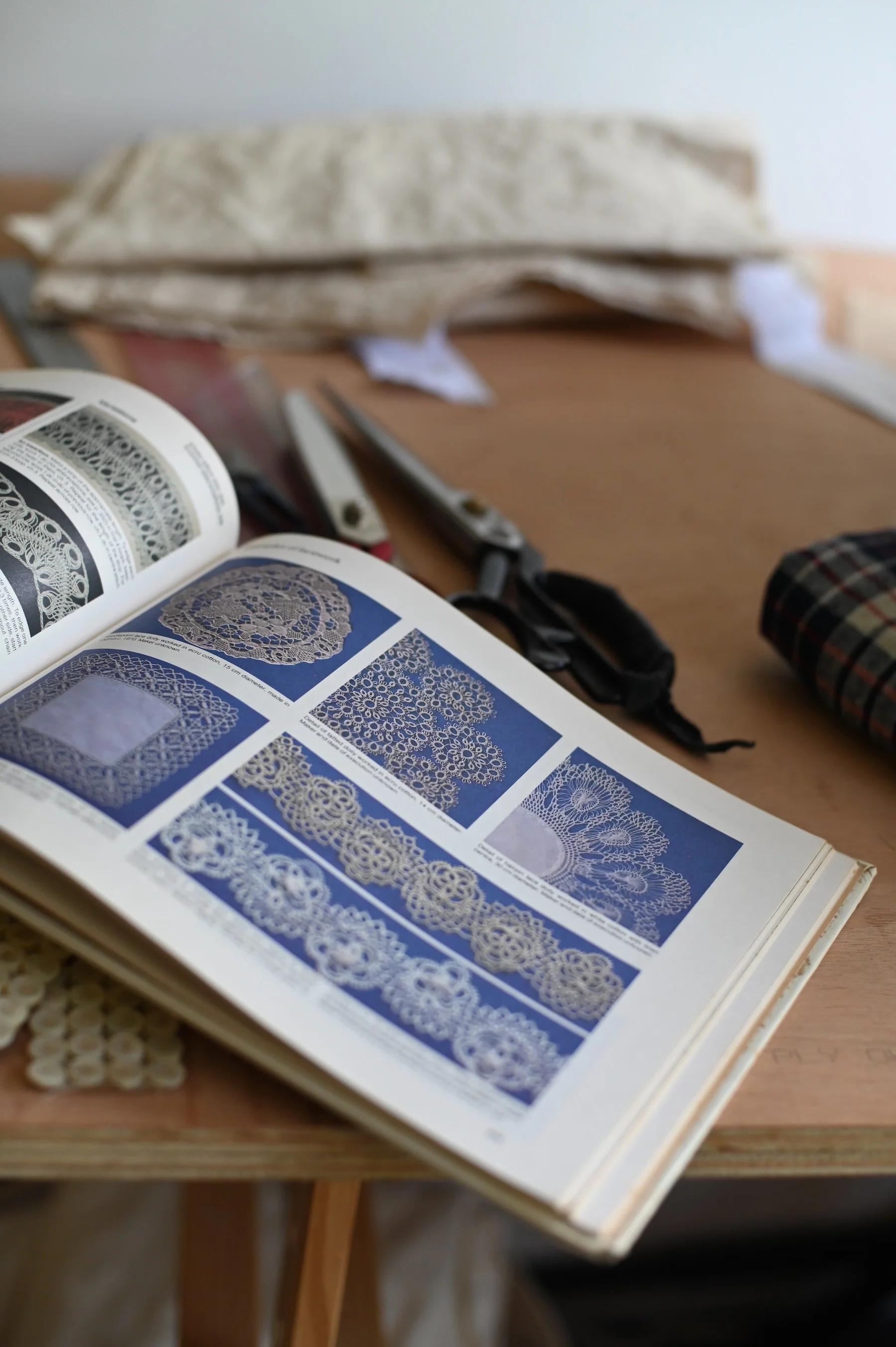 Open book with lace patterns on wooden table. Sewing tools and fabric pieces nearby.