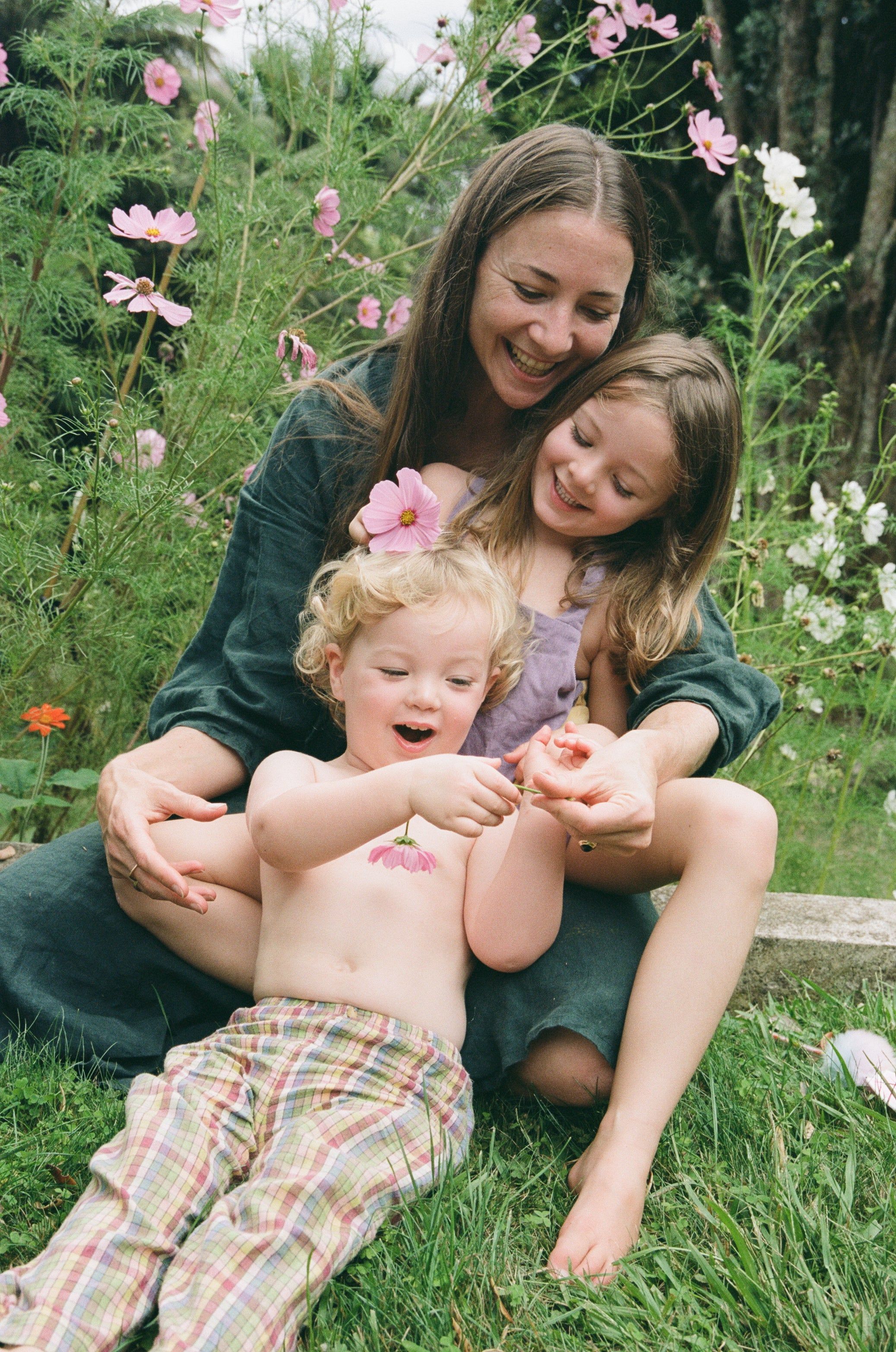 Woman on grass holding two children. Children playing with pink flowers. All smiling.