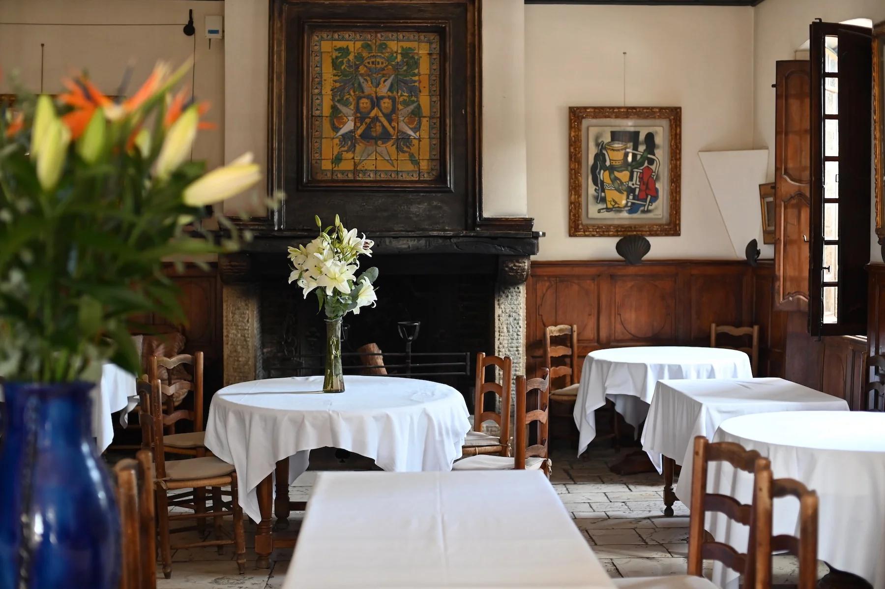 Cozy café with wooden chairs and white tablecloths. White lilies on central table, paintings on walls, and fireplace with large artwork above.