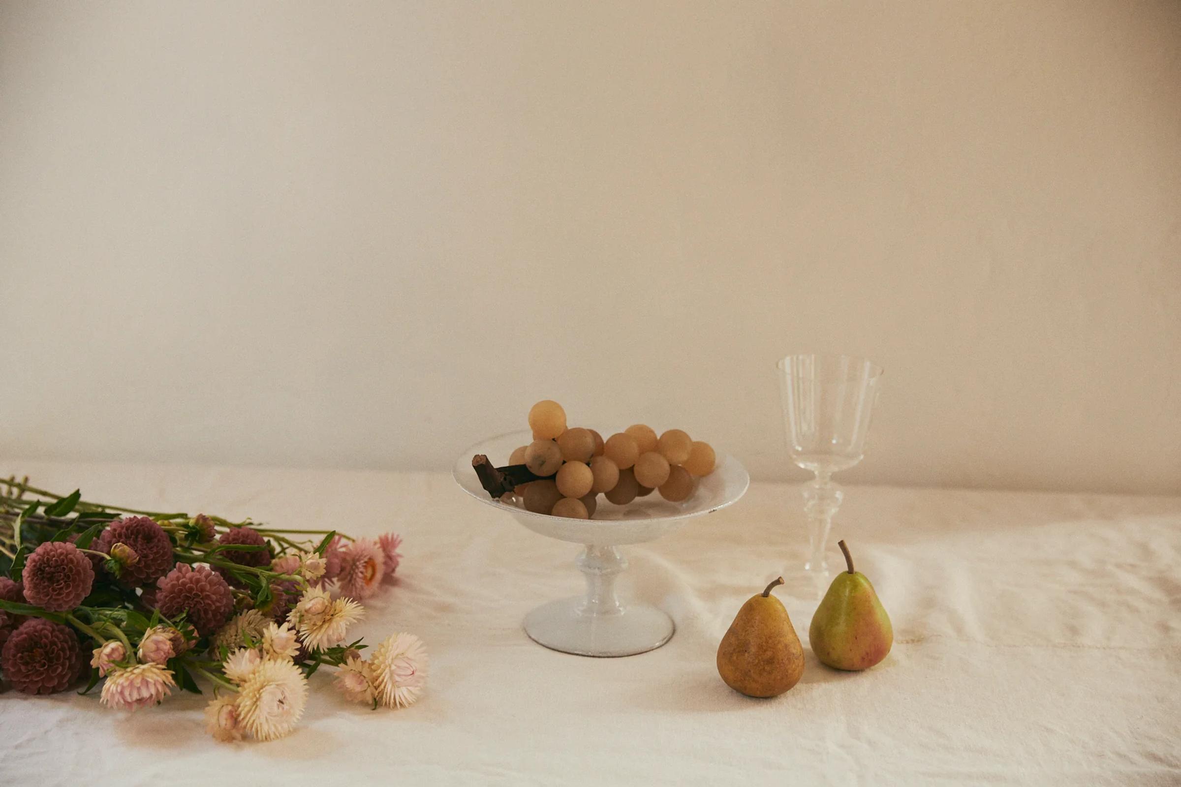White pedestal bowl with carved grapes and pears, beside wine glass and pink flower bouquet on light tablecloth. Plain, light-coloured wall background.