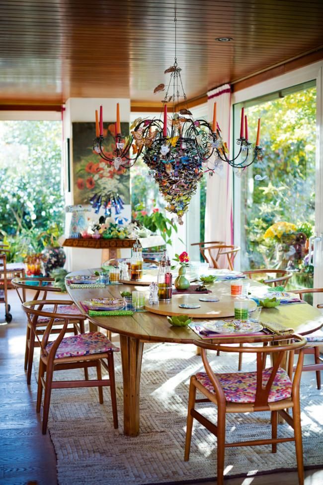 Dining area with wooden table and colourful chairs. Vibrant chandelier above. Large windows show lush greenery outside.