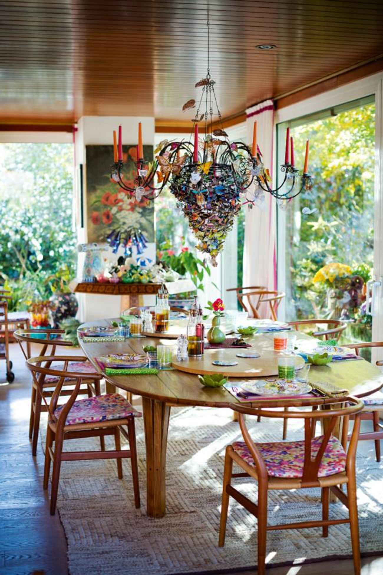 Dining area with wooden table and colourful chairs. Vibrant chandelier above. Large windows show lush greenery outside.