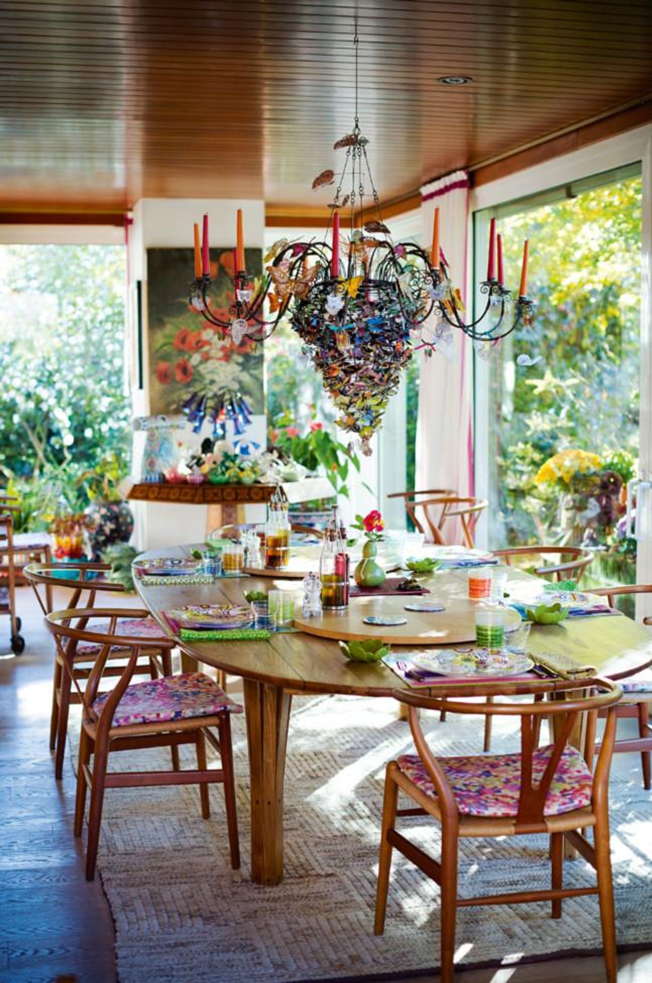 Dining area with wooden table and colourful chairs. Vibrant chandelier above. Large windows show lush greenery outside.