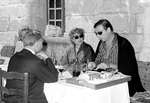 Black and white photo of three people at outdoor restaurant table. Wine glasses, water bottle visible. Stone walls in background.