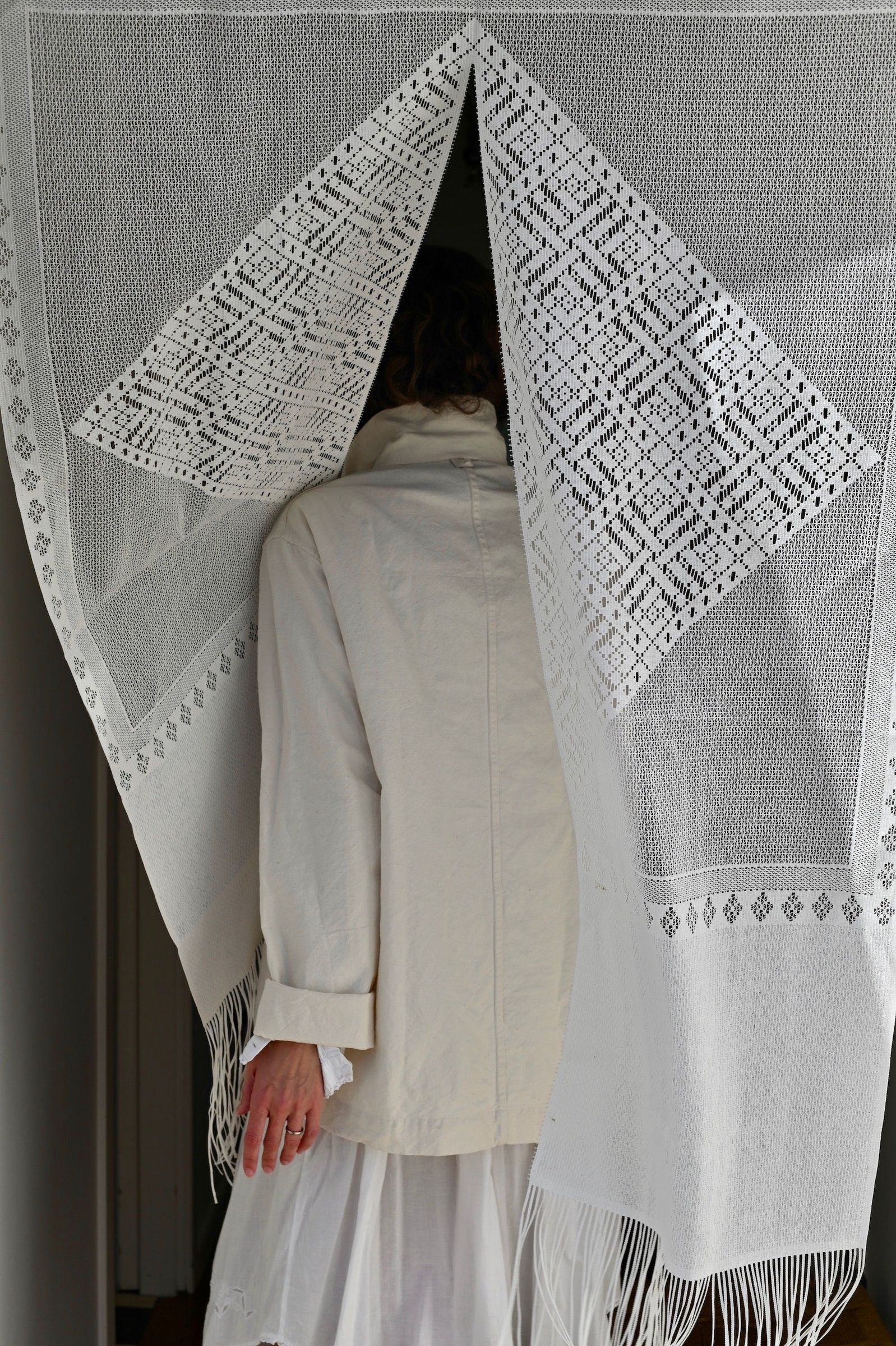 Person in light coat and skirt walking through doorway with lace curtain. Hand touches curtain, showcasing Oliver Church's practical design.