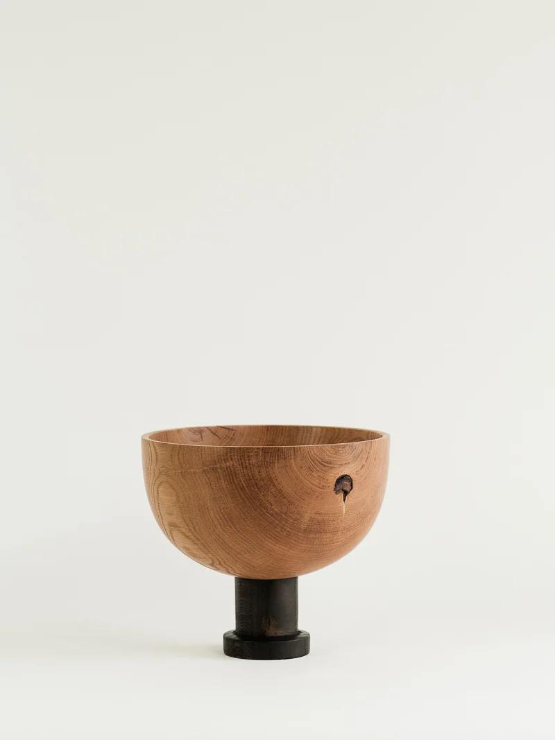 A handcrafted wooden bowl with a smooth, natural grain finish rests on a black cylindrical pedestal. Featuring Titirangi design, a small hole near the rim adds a rustic touch. The background is light and neutral.