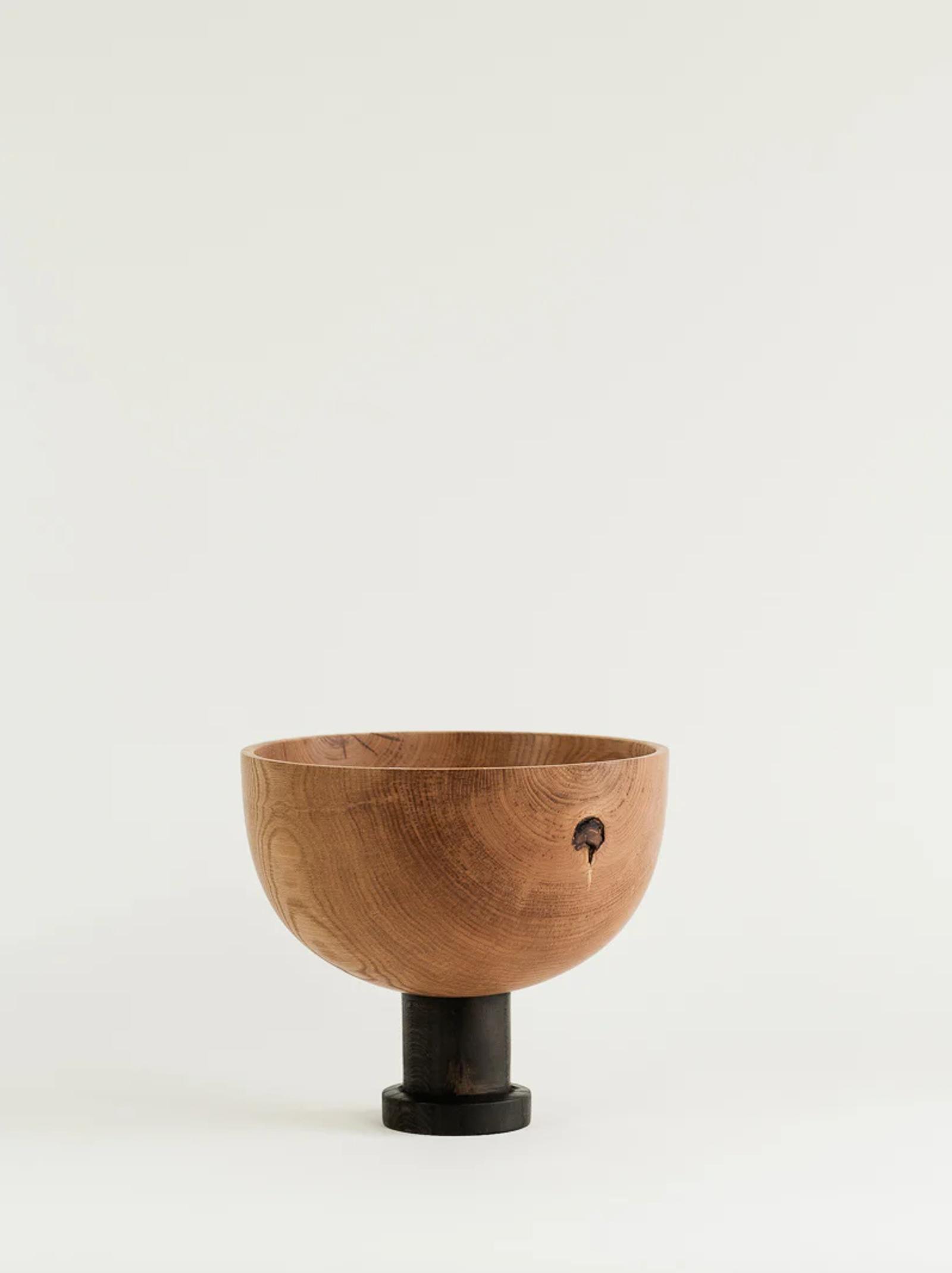 A handcrafted wooden bowl with a smooth, natural grain finish rests on a black cylindrical pedestal. Featuring Titirangi design, a small hole near the rim adds a rustic touch. The background is light and neutral.