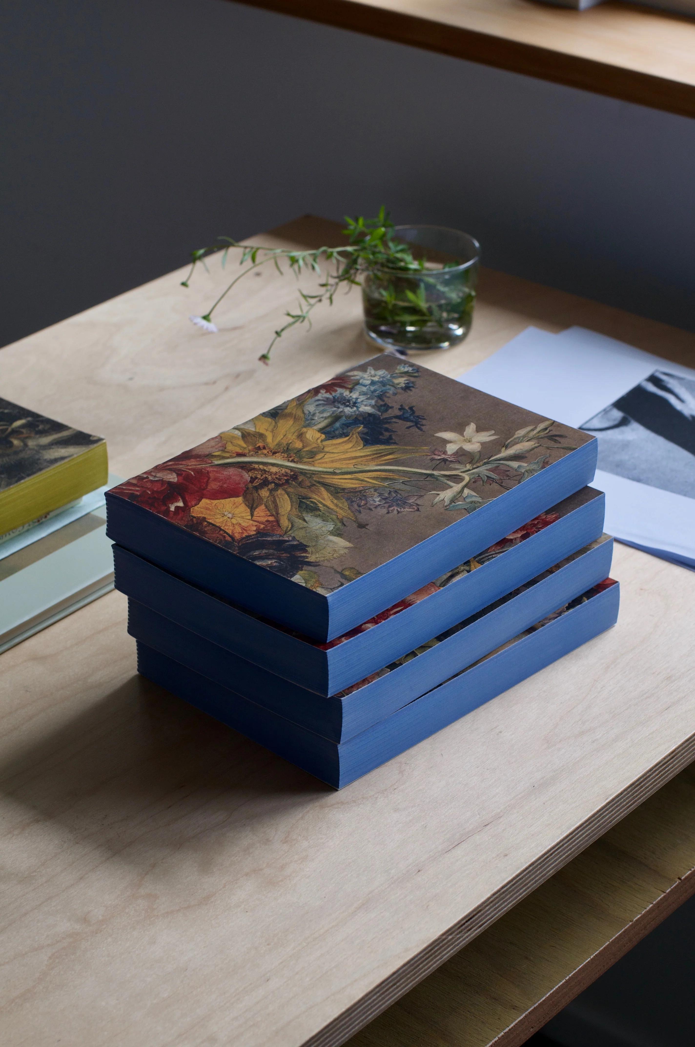 Four blue-edged books with floral covers on wooden table. Glass with small plant, papers, and yellow-edged book nearby. Natural light illuminates scene.