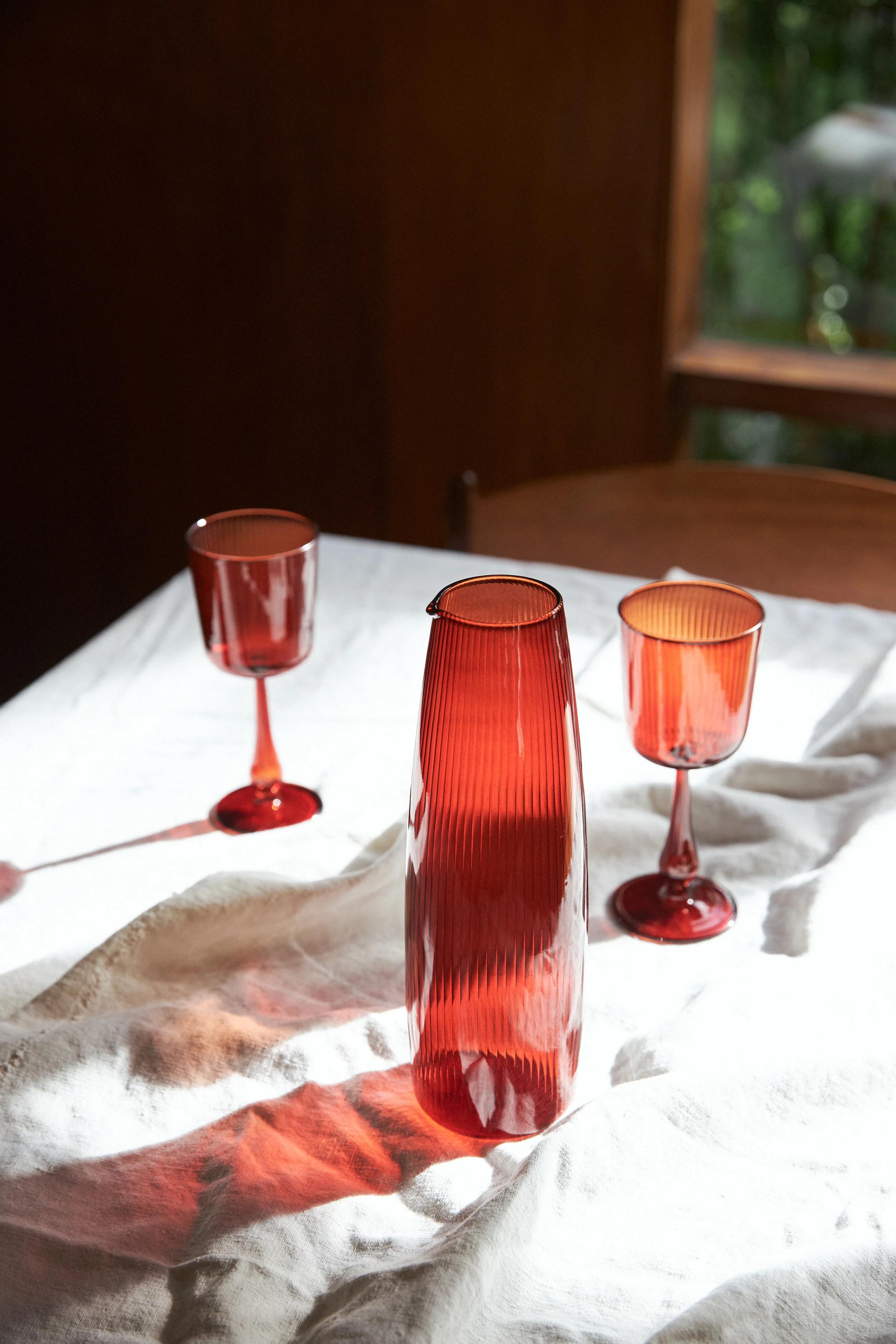 A ribbed red glass carafe and matching wine glasses on a white tablecloth. Sunlight creates reflections from the glassware on the fabric.