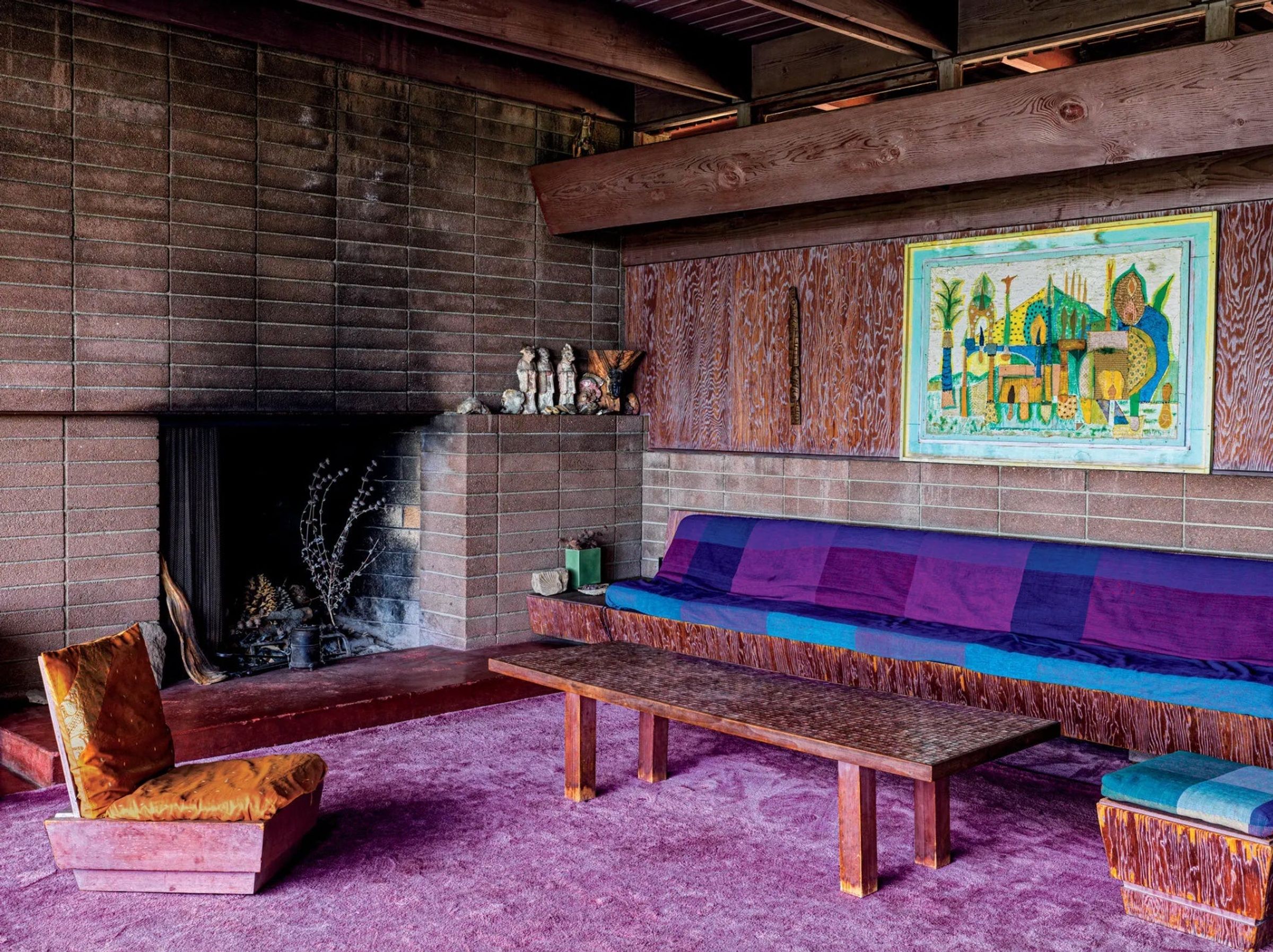 A cozy living room with large brick fireplace, wooden walls, purple carpet, a large purple and blue couch, coffee table and orange chair. There's a bright painting and sculptures on the mantle.