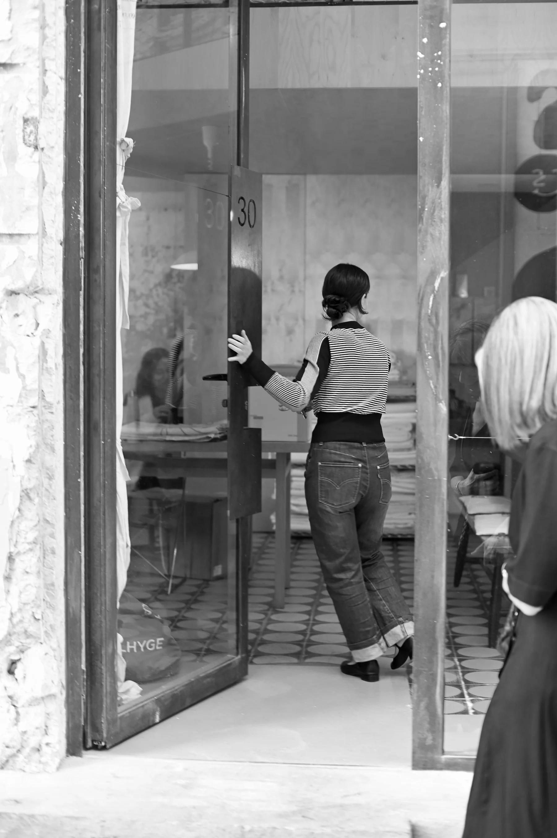 Woman in jeans and striped top holds open shop door. Another person with light hair stands in front of door.