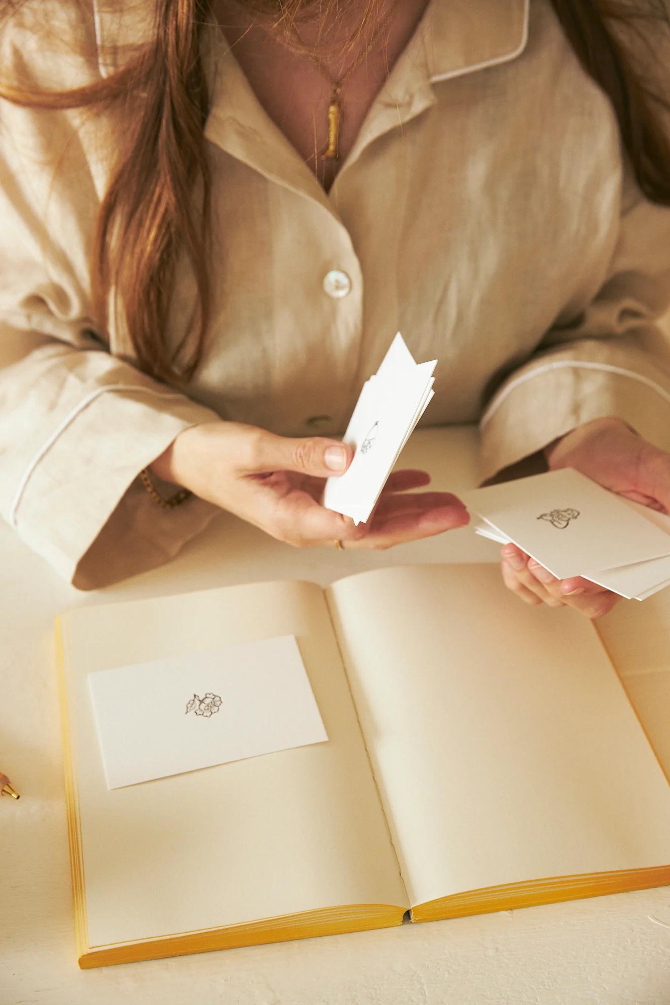 Person in beige shirt sorting through cards with drawings. Open notebook on table.