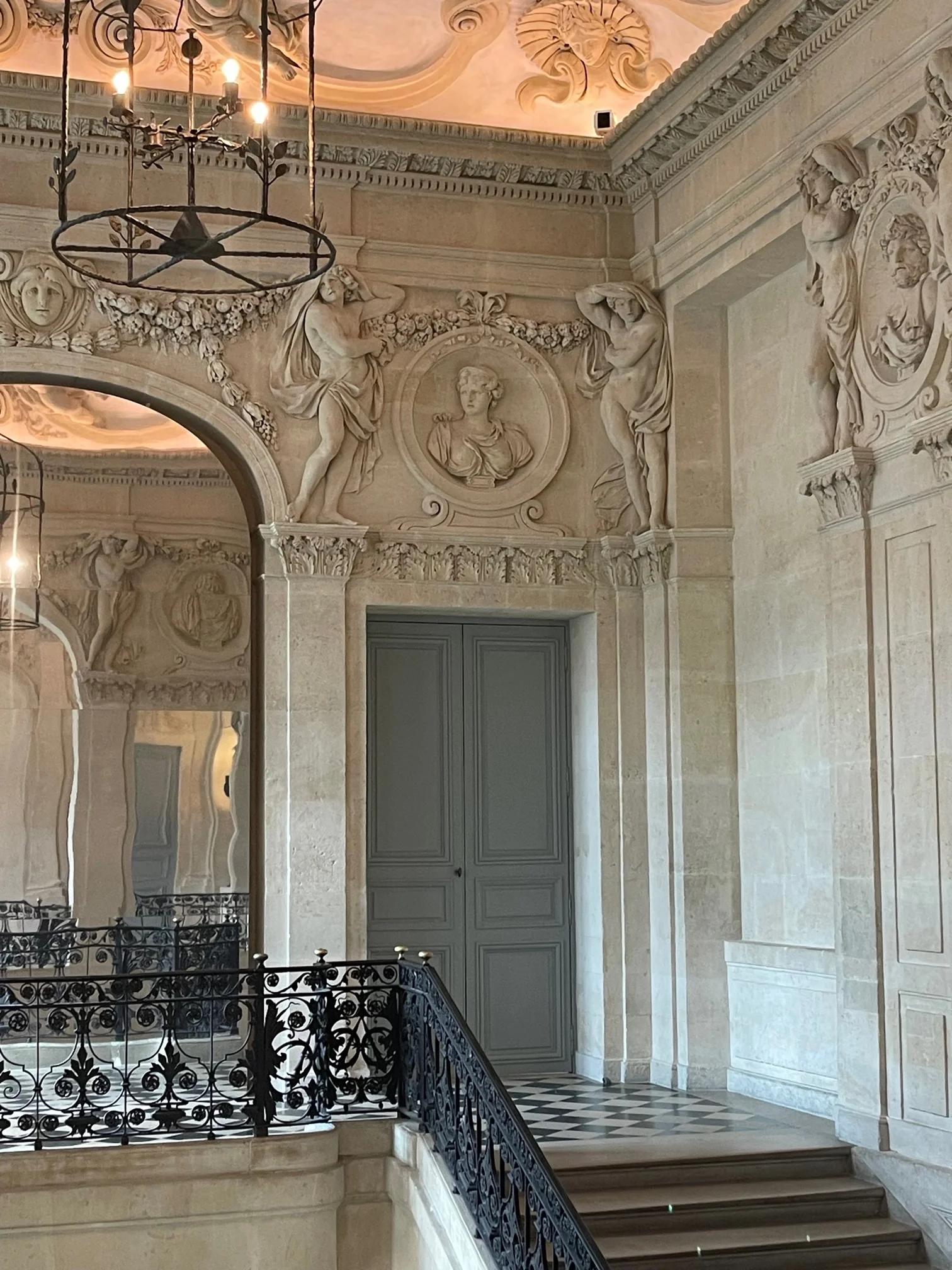 Grand staircase in ornate building. Sculptures on walls and arches. Black wrought-iron handrail. Large grey door in background.