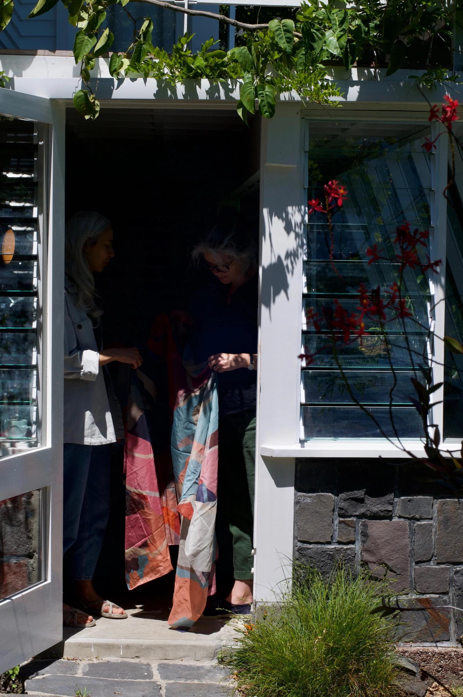 Two people in doorway examining geometric quilt. Sunlit greenery and red flowers in foreground.