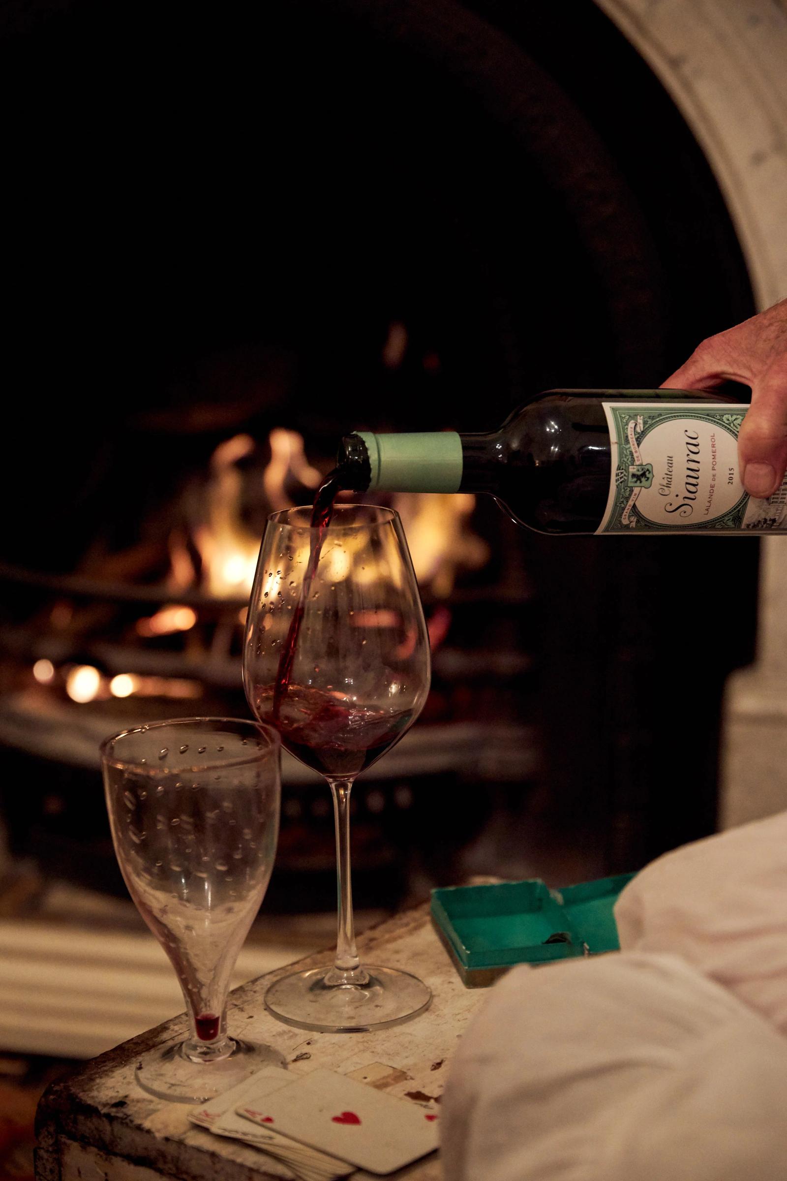 A hand is pouring red wine from a bottle into a glass next to another empty glass, placed on a wooden table. A fire burns in the background.