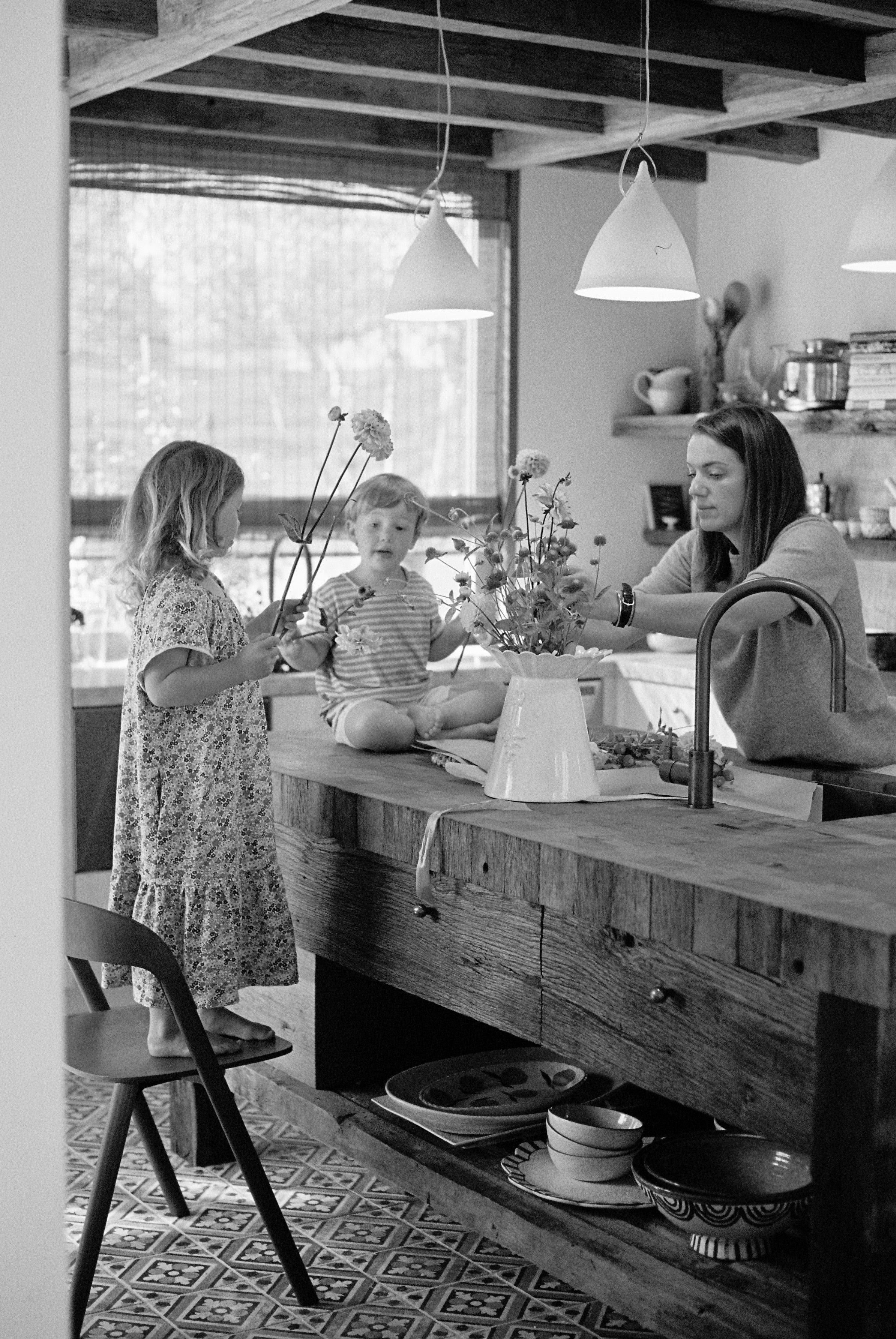 Black and white image of woman and two children in rustic kitchen. Woman arranges flowers, one child on island, another holding flower.