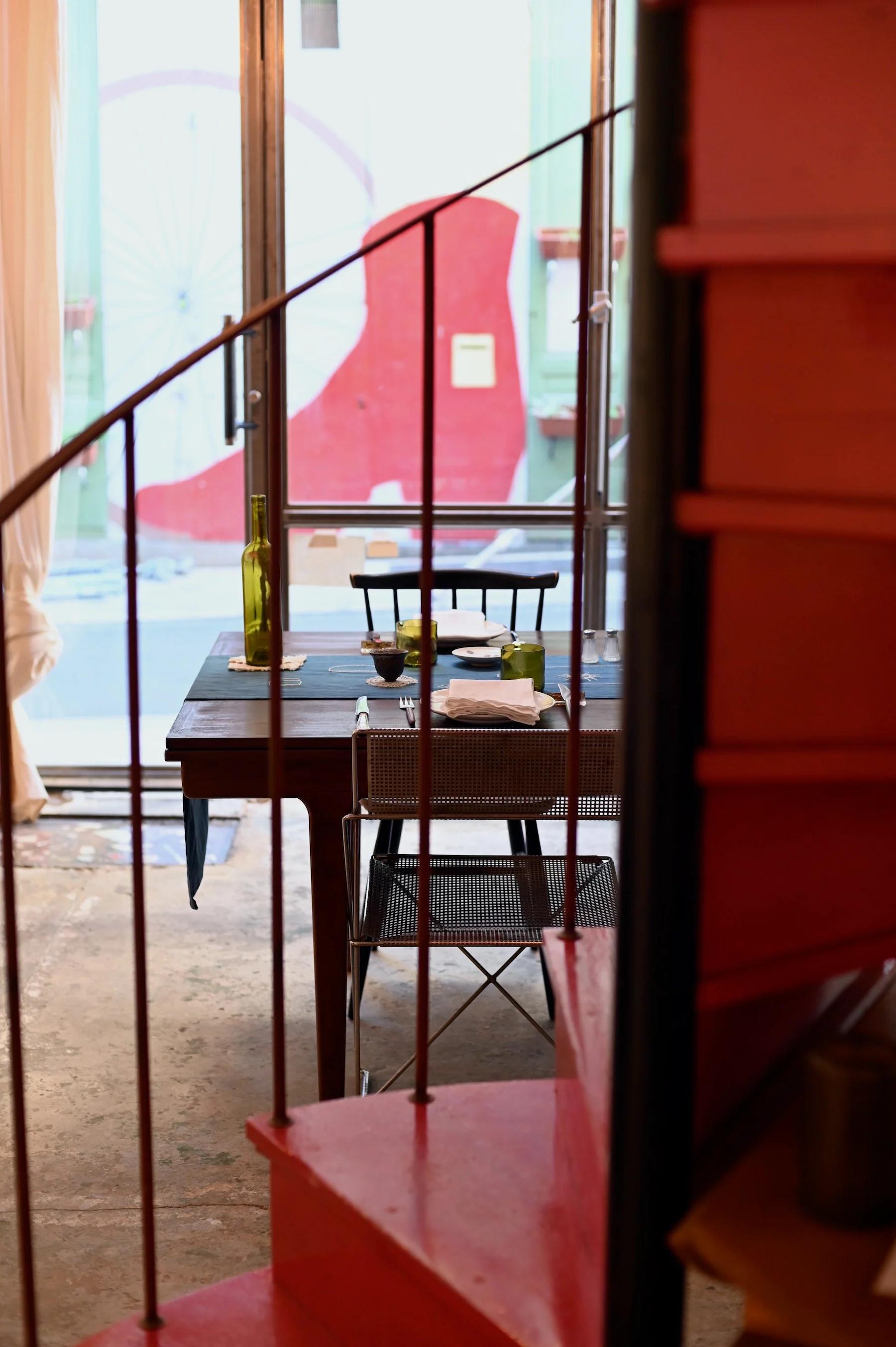 Dining table with embroidered linens near large window. Red wall visible outside. Red spiral staircase in foreground.