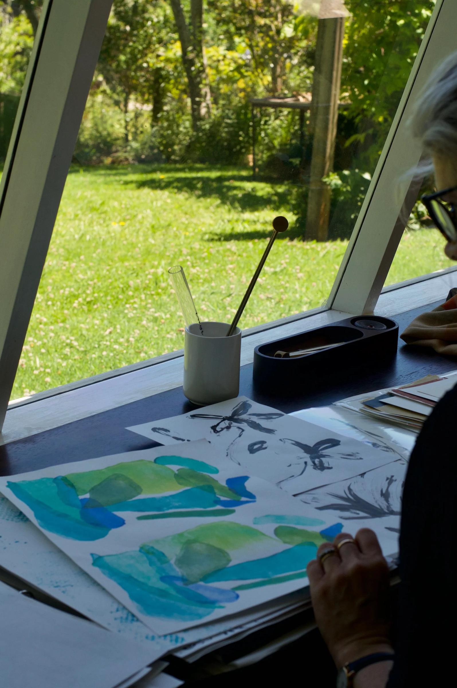 Person painting at desk by large window. Art supplies scattered. View of lush garden outside.