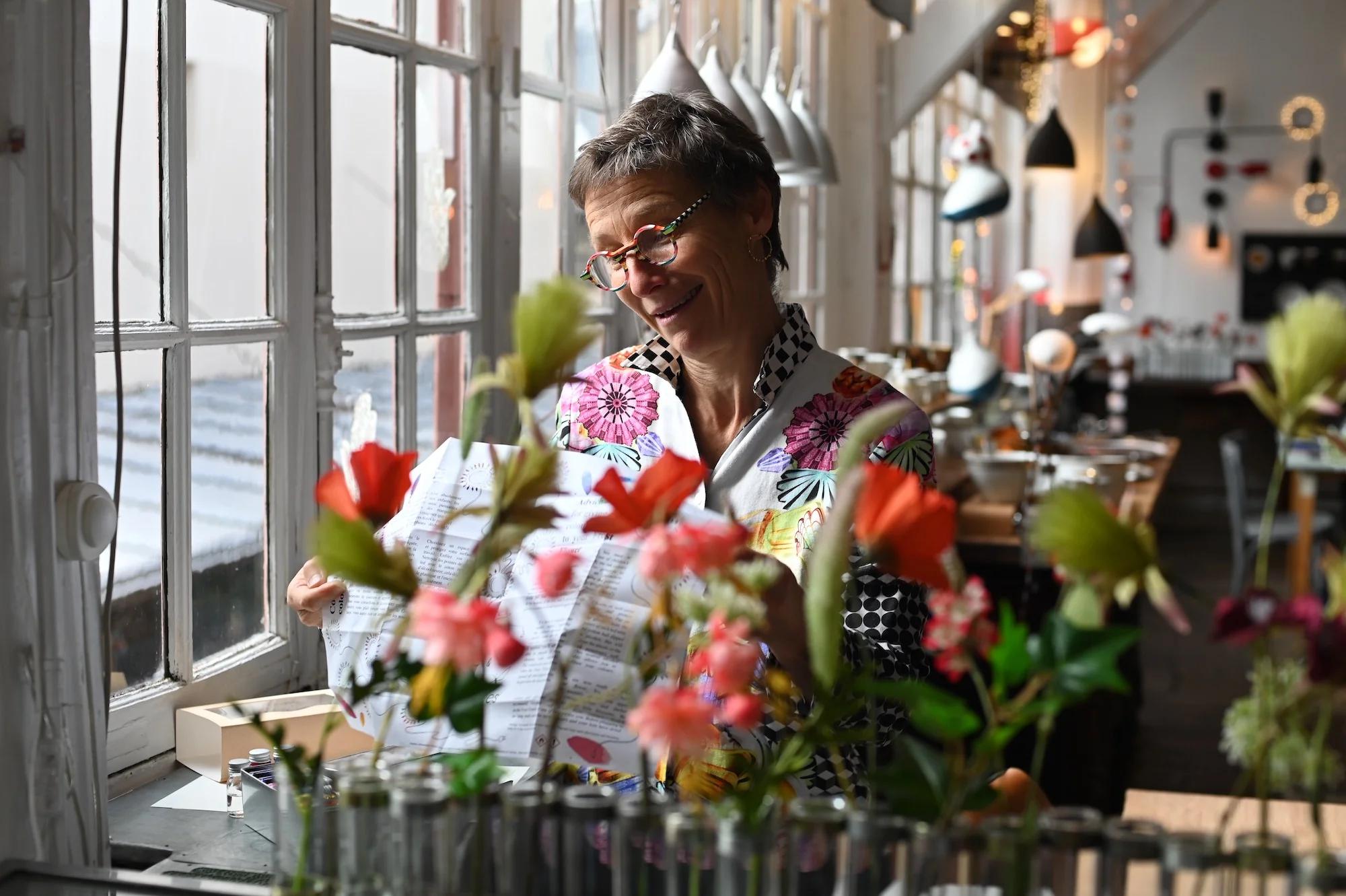 Short-haired person with glasses smiling at paper. Room has flowers, hanging lamps, and colourful decor. Eclectic shirt.