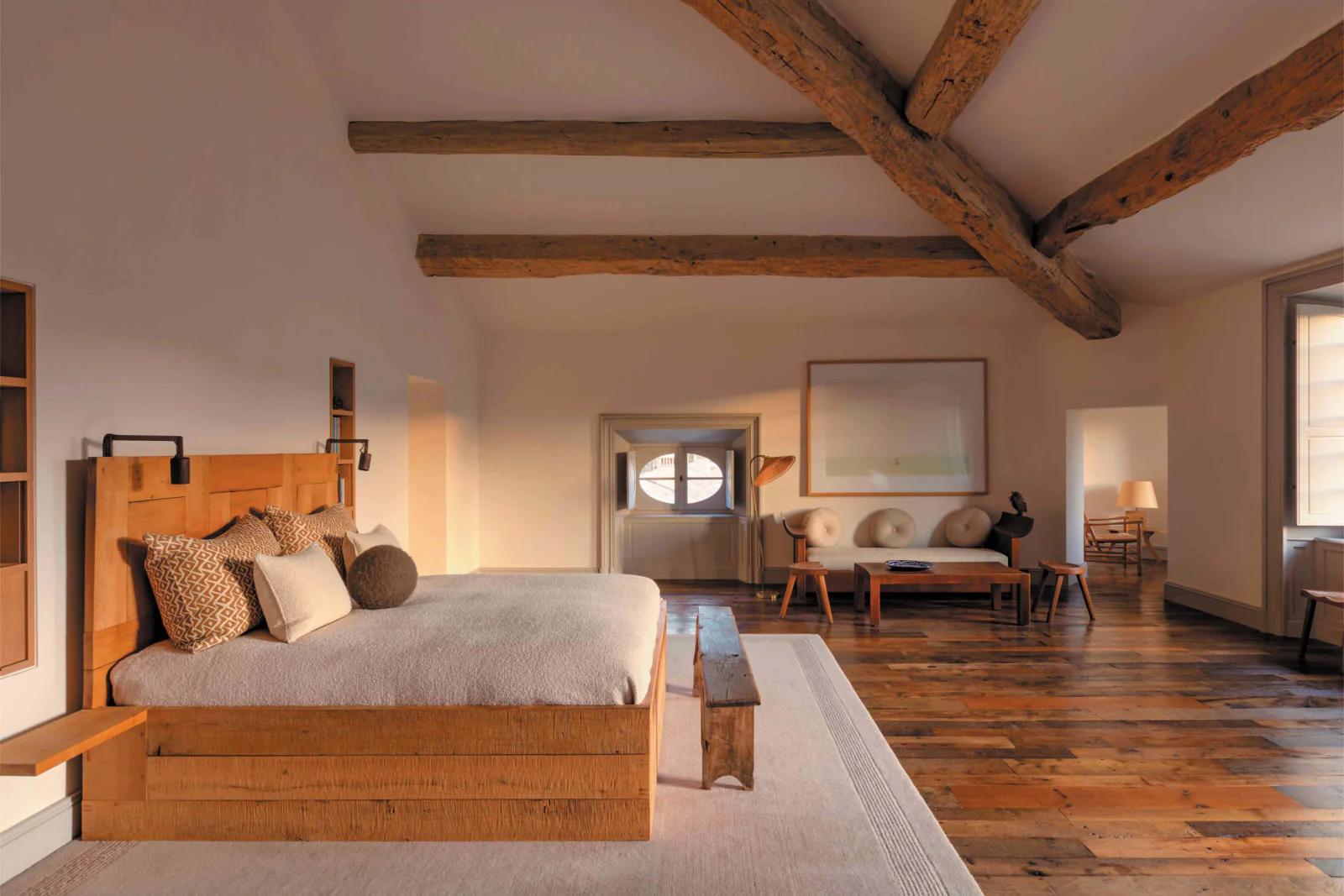 A rustic loft bedroom with exposed beams and large windows. It features a wooden bed, bench, and cozy seating area, echoing Provençal architecture.