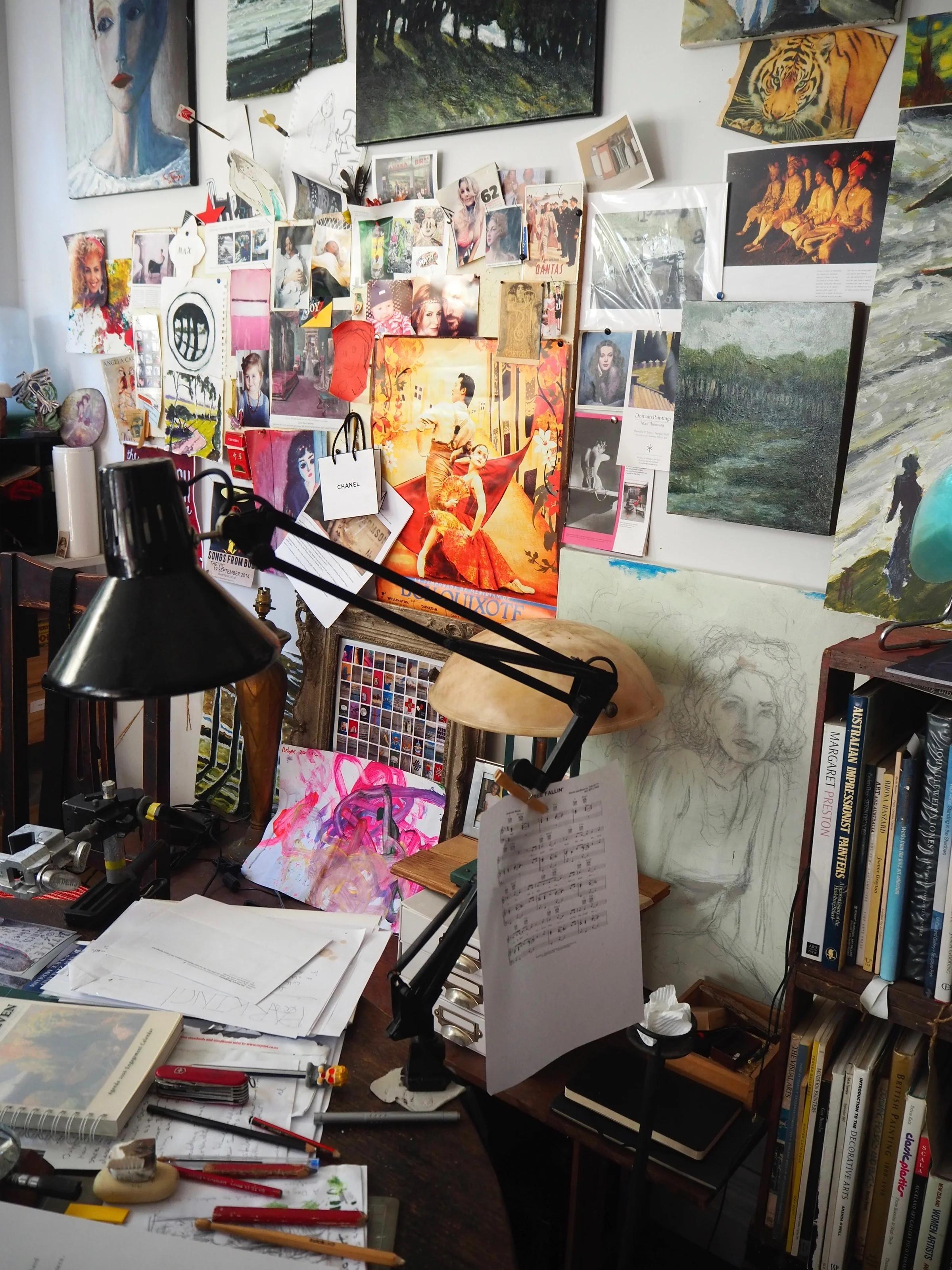 Dark desk lamp on cluttered table. Max Thomson's paintings and sketches cover wall. Book stacks on shelves to right.