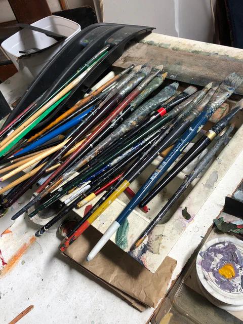 Paintbrushes with stained handles on messy, paint-splattered surface. Art supplies and palette nearby. Max Thomson's workspace.