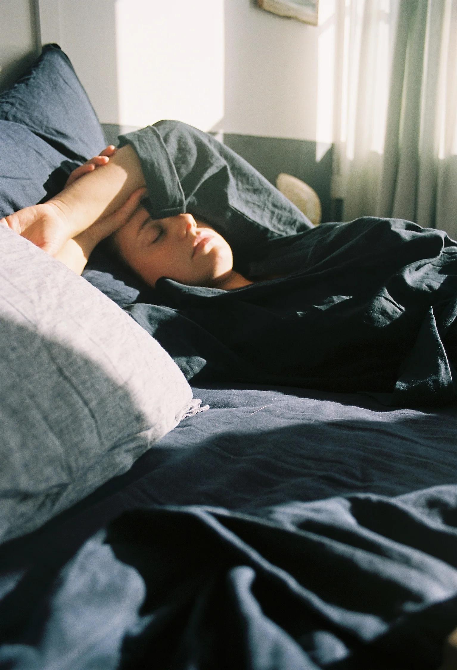 A person lying on a bed covered by dark blue sheets, resting with eyes closed, as sunlight filters through a window, evoking a relaxed seaside mood.