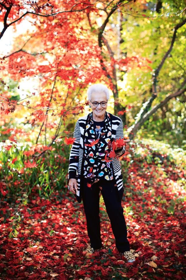 Elderly woman in striped cardigan and patterned blouse amid autumn foliage. Holds red leaves, smiling.