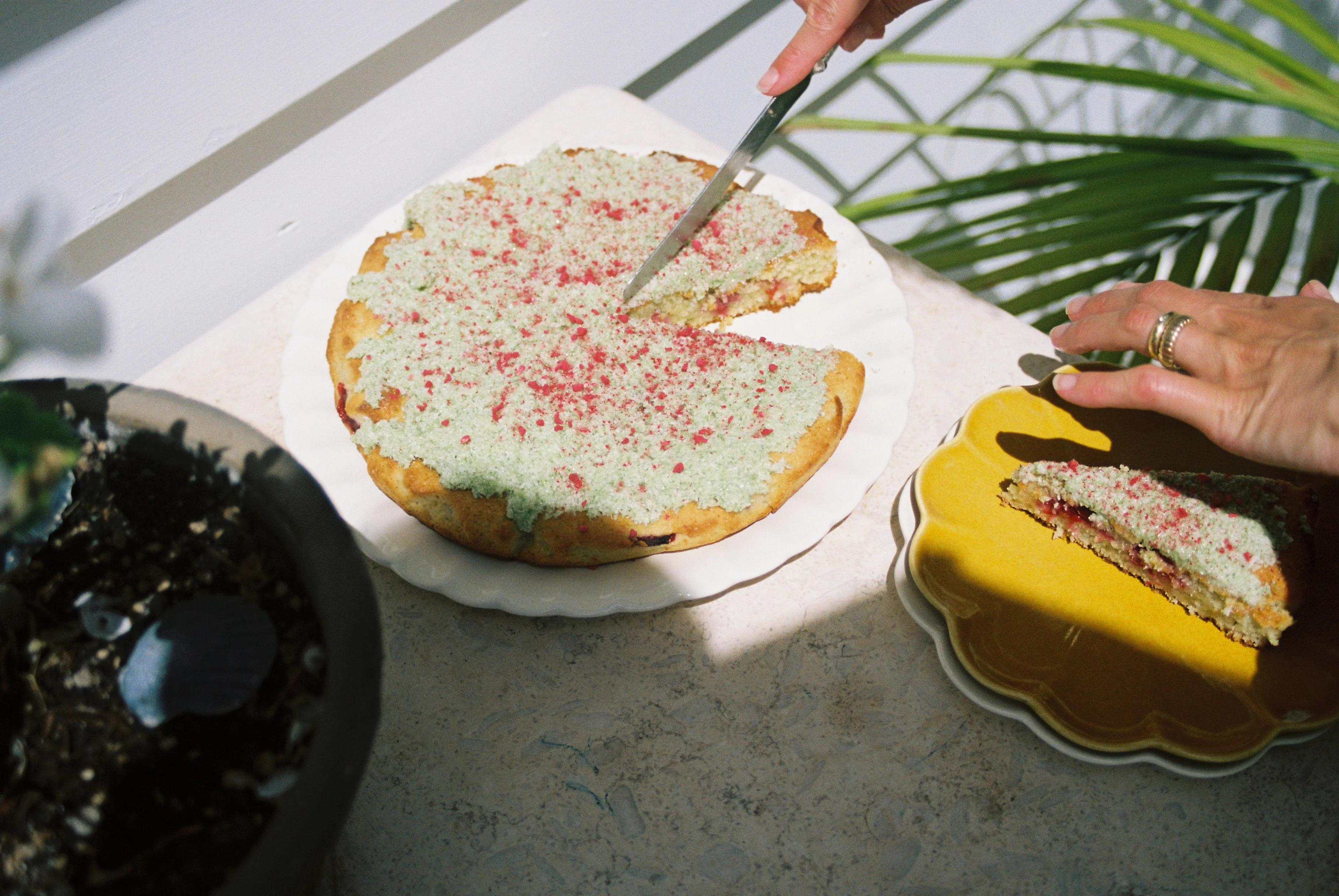 Person cutting sprinkle-covered cake. Slice transferred to yellow plate. Potted plant and sunlight in cosy setting.