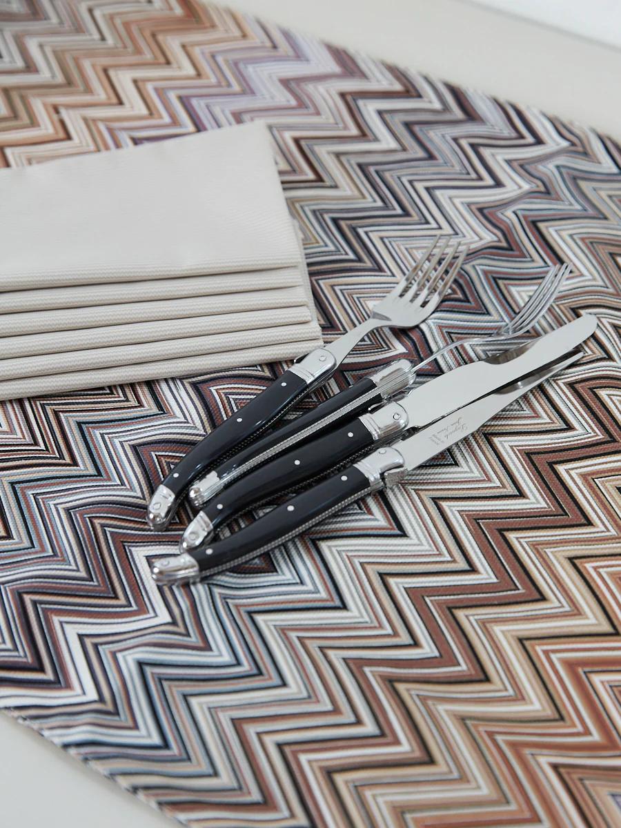 Three black-handled utensils on zigzag placemat. Brown, gray, white shades. Folded white napkins nearby.