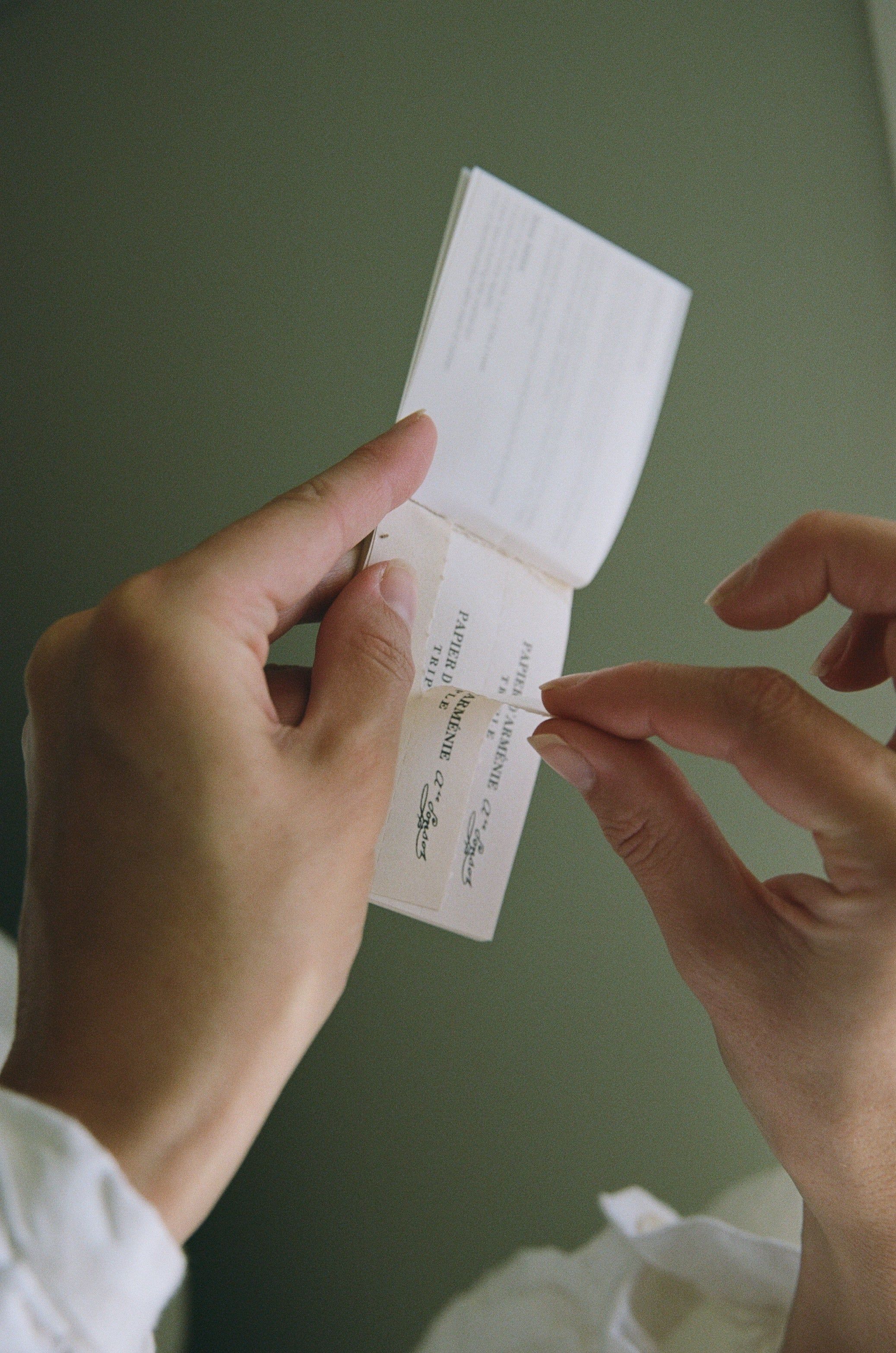 Close-up of hands with manicured nails opening a small booklet with perforated, scented pages. The muted green background adds a touch of serenity.