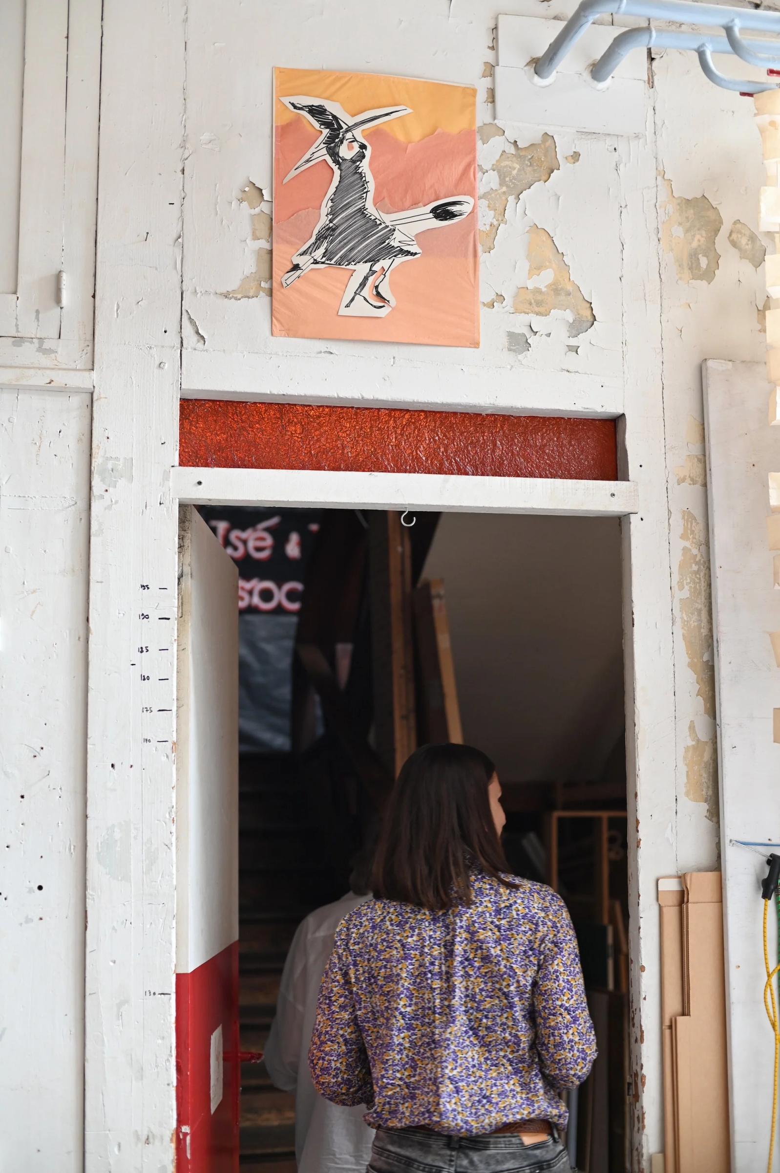 Long-haired person in floral shirt and jeans in doorway. Framed image above of woman with ladle and knife. Worn, partially white wall.