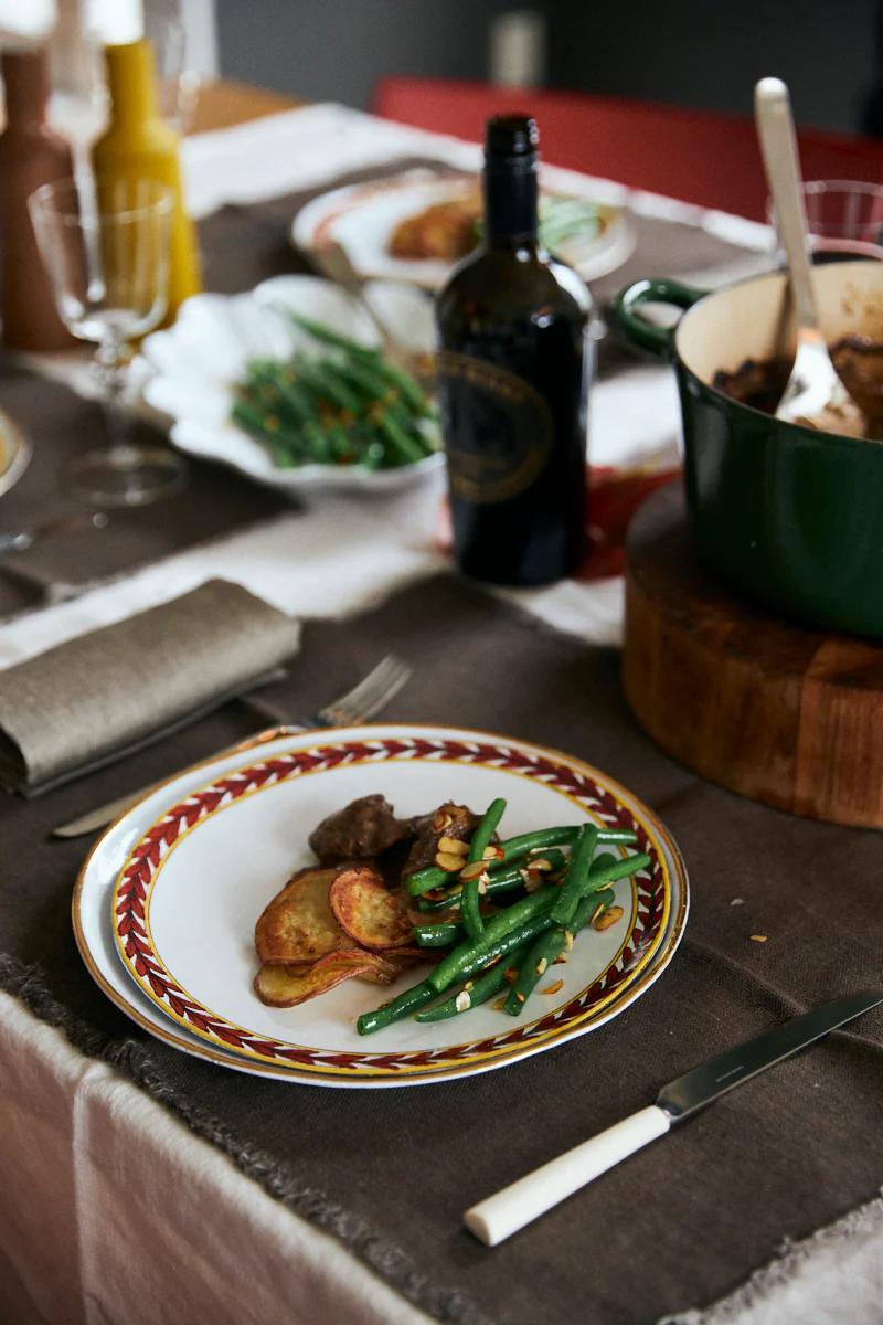 Colourful plate with green beans, roasted potatoes, and beef casserole. Black bottle, green pot, glassware on grey tablecloth.