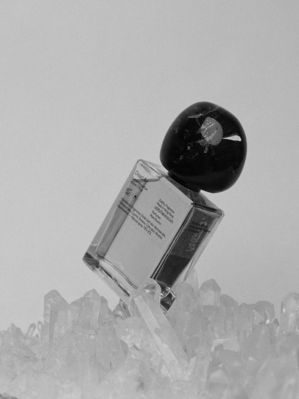 Black and white image of perfume bottle with round, dark cap on crystals. Label with text visible.