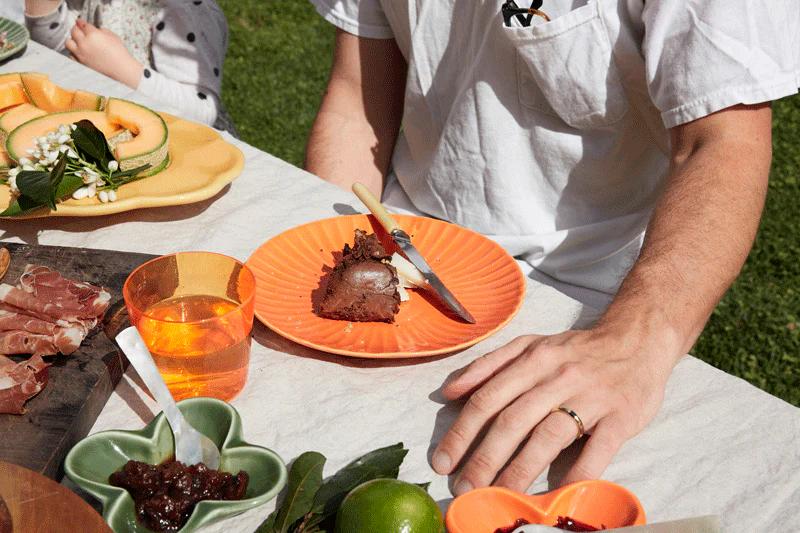 Close-up of person at outdoor table with cake slice on orange plate. Table set with fruit, jam, and meats. Person wears white shirt and wedding ring.