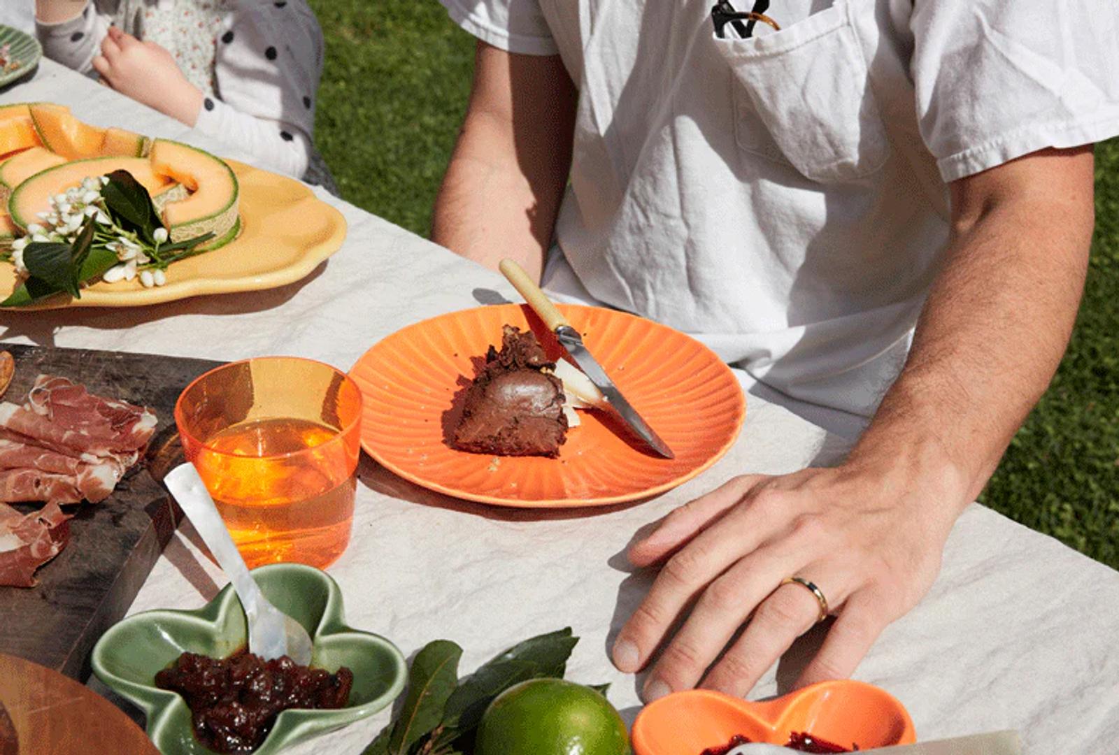 Close-up of person at outdoor table with cake slice on orange plate. Table set with fruit, jam, and meats. Person wears white shirt and wedding ring.