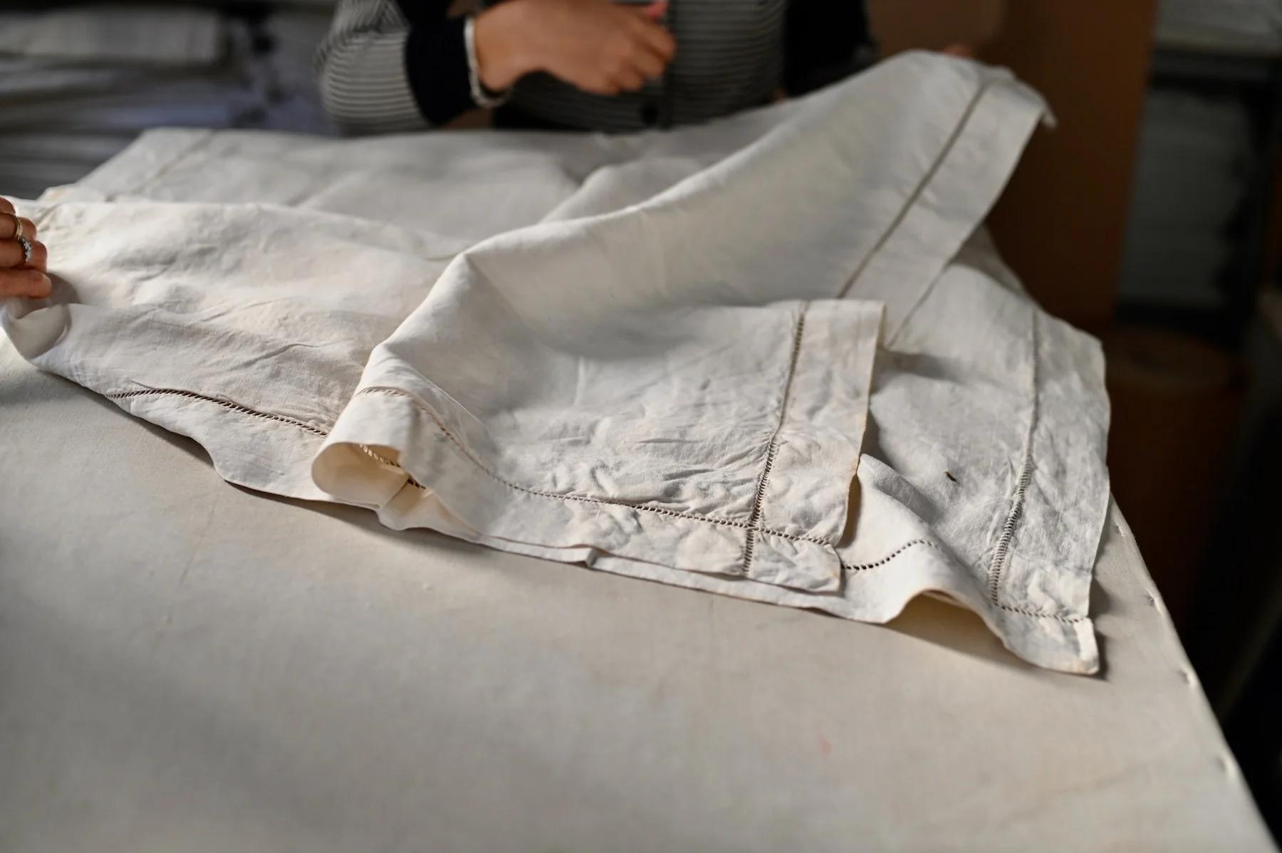 Person folds large beige fabric with visible stitching. Hands barely visible in background.