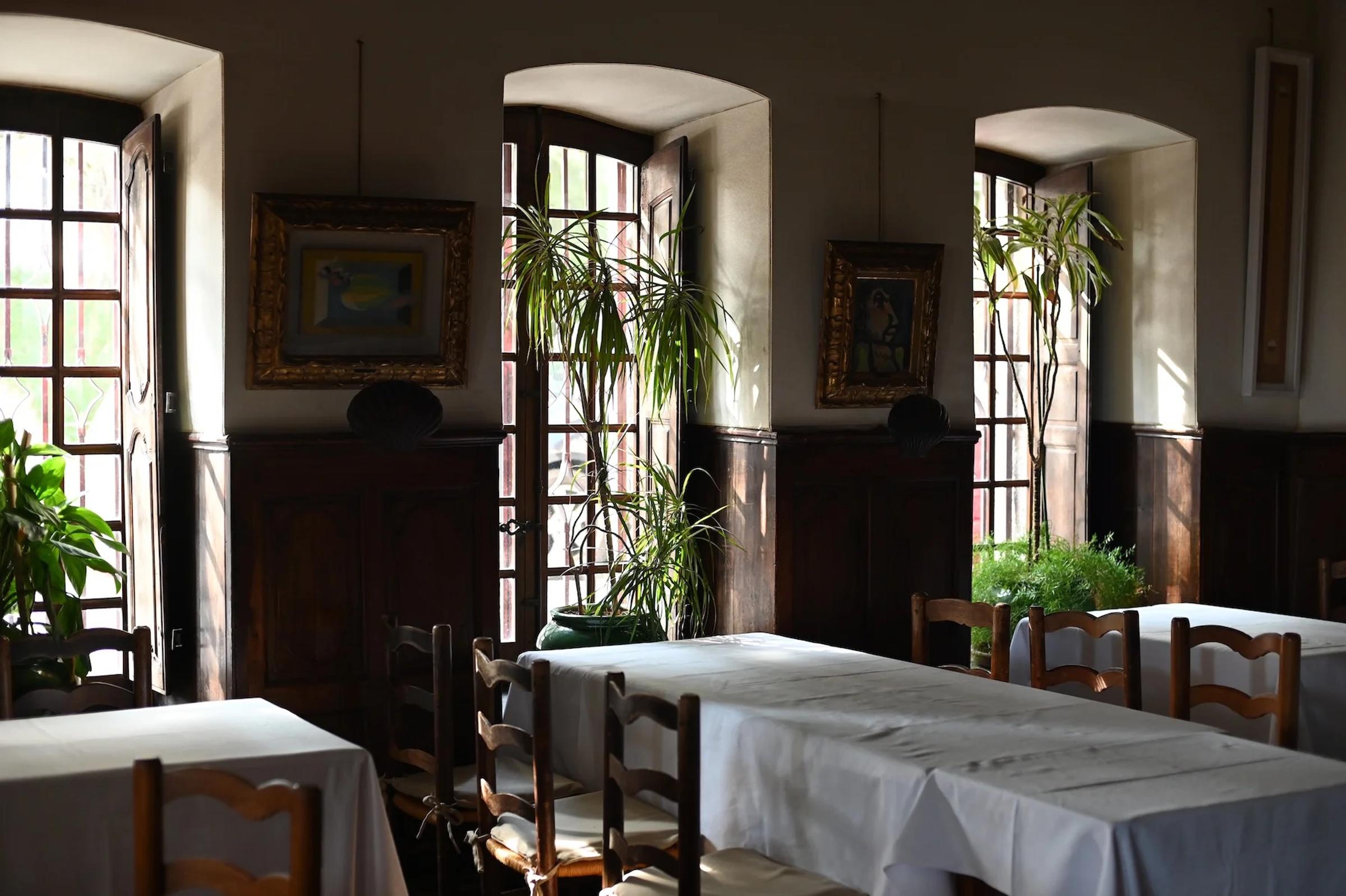 Sunlit restaurant with wooden chairs and white-clothed tables. Large windows illuminate space, showcasing plants and framed paintings.